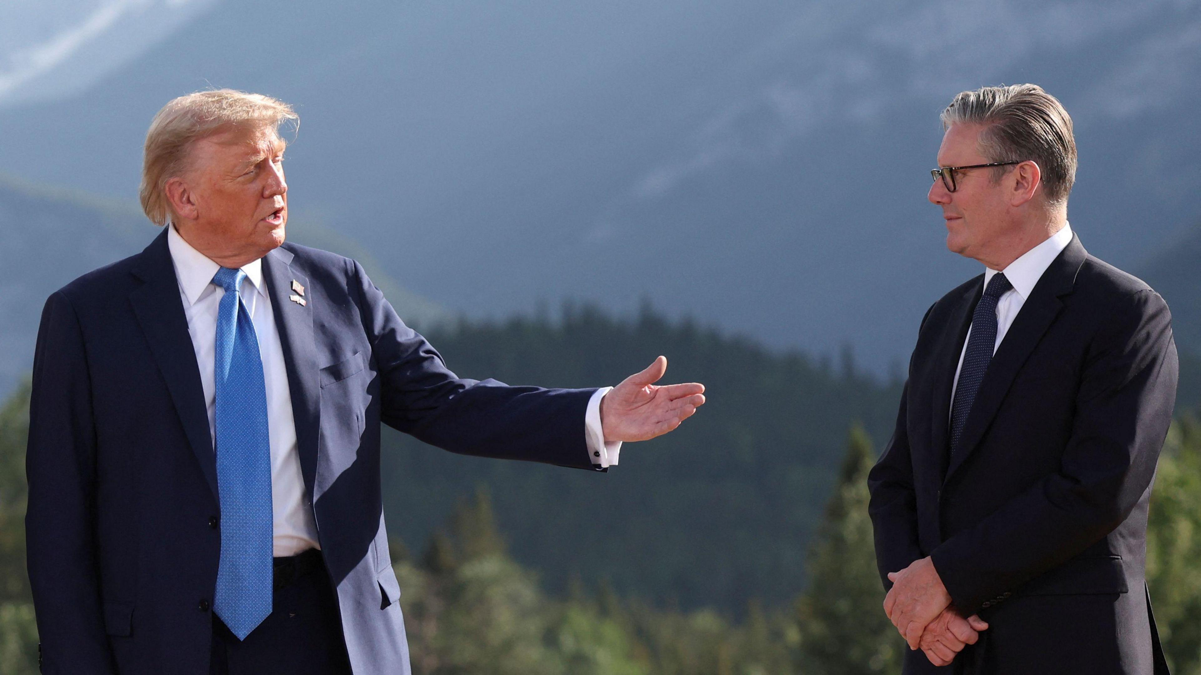 President Donald Trump gestures towards British Prime Minister Keir Starmer with his outstretched left hand. He is wearing a navy suit, white shirt and light blue tie. Sir Keir, who is standing with his arms folded is wearing a black suit, white shirt and navy tie. The leaders are standing in front of the Canadian Rockies. Lush green forests can be seen in the background