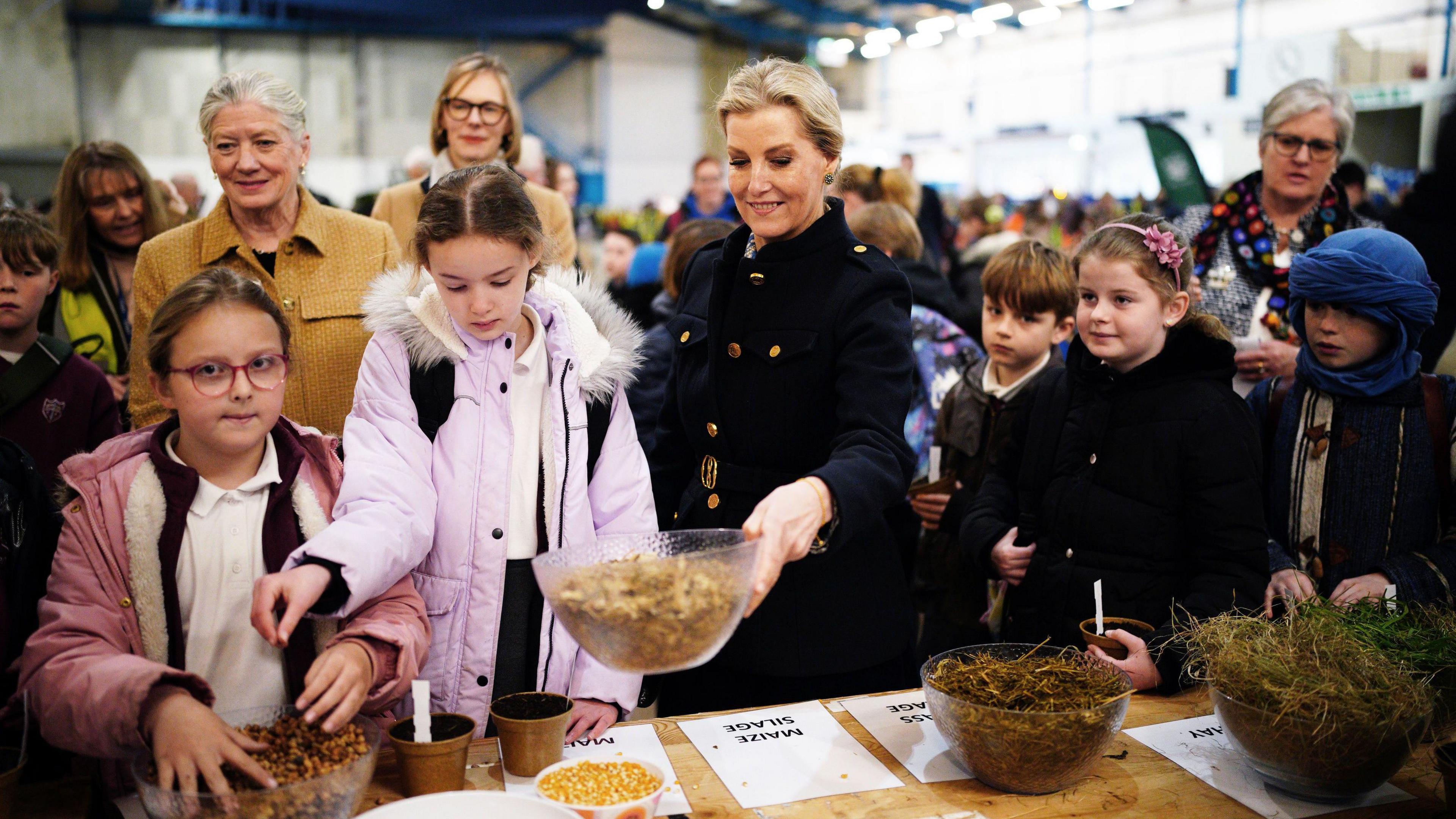 Duchess of Edinburgh looking at bowls of maize and grass, alongside schoolchildren