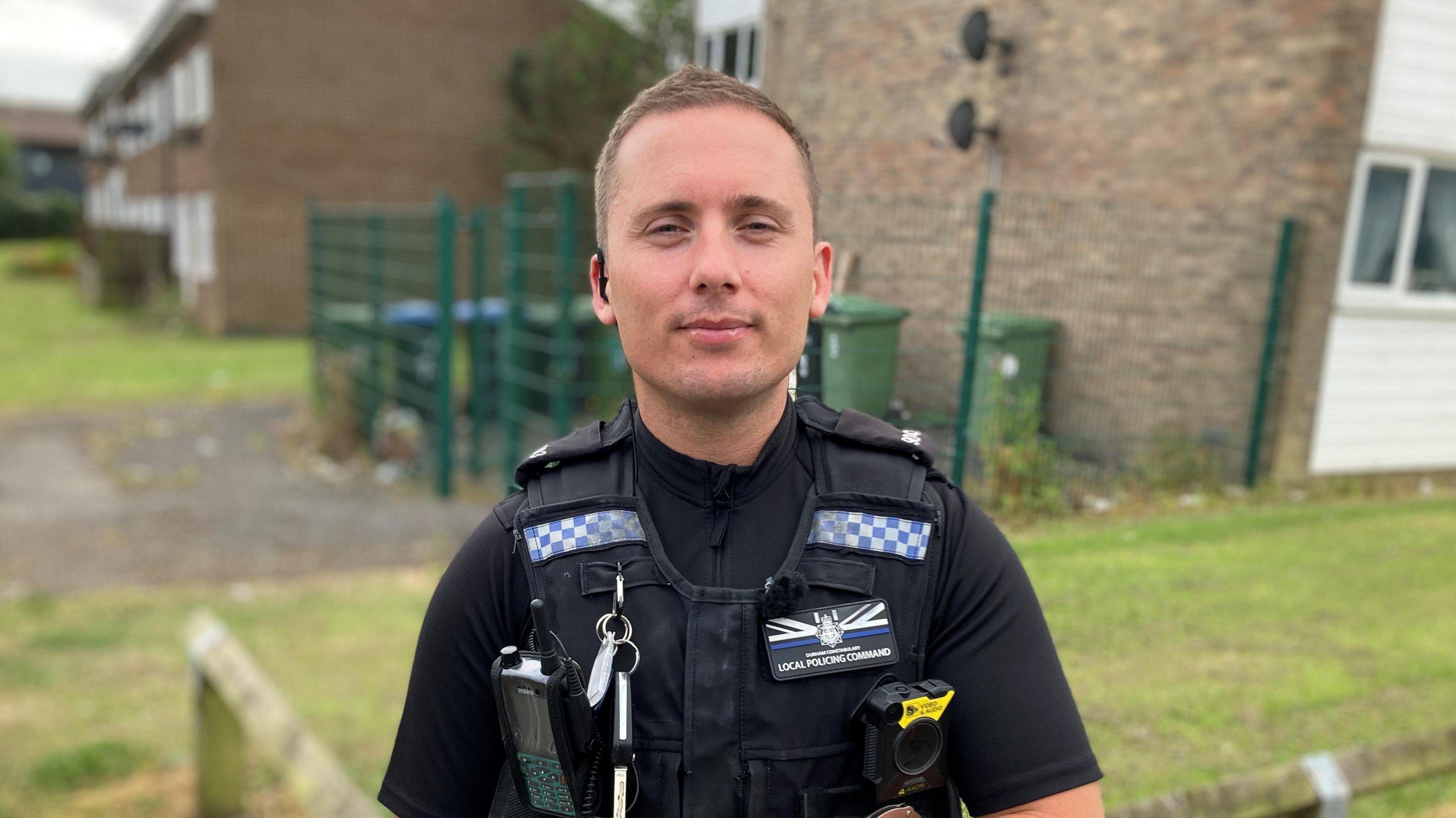 PC Furlonger, who is wearing his uniform and standing in a housing estate. He has short cropped light brown hair and is smiling.