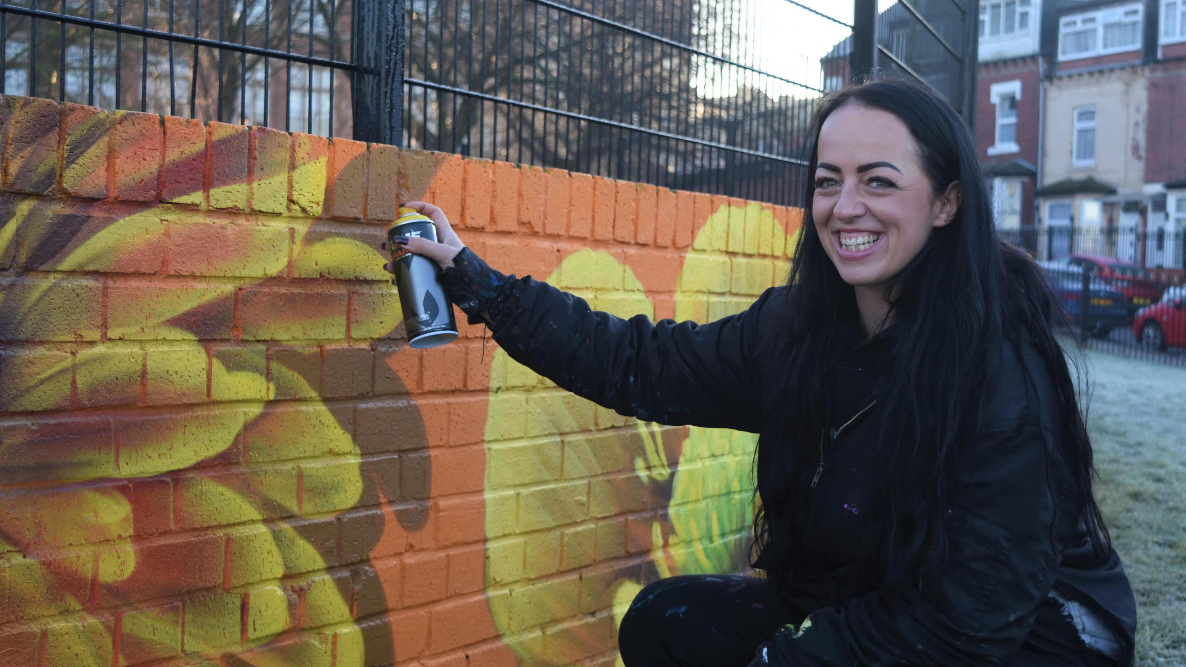 A smiling woman with long black hair crouches and poses with a spray can pointed towards the brightly-coloured mural.