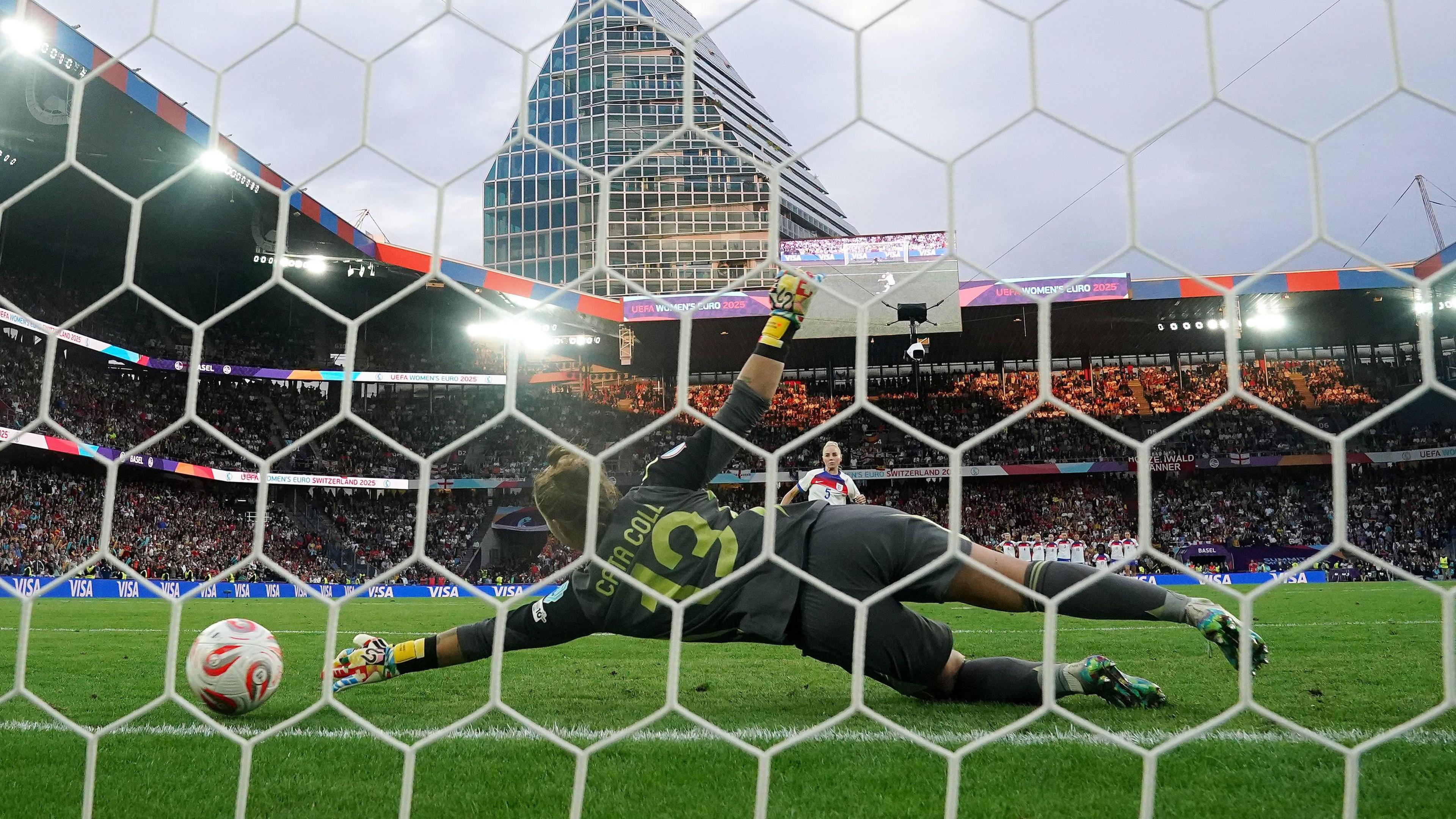 a photo taken through a goal net. In the foreground, a keeper in dark green kit dives to her left, unable to make contact with a football. In the background, the top half of a footballer in an England shirt is visible. Behind her are packed stands on all sides, and a large skyscraper in the background.