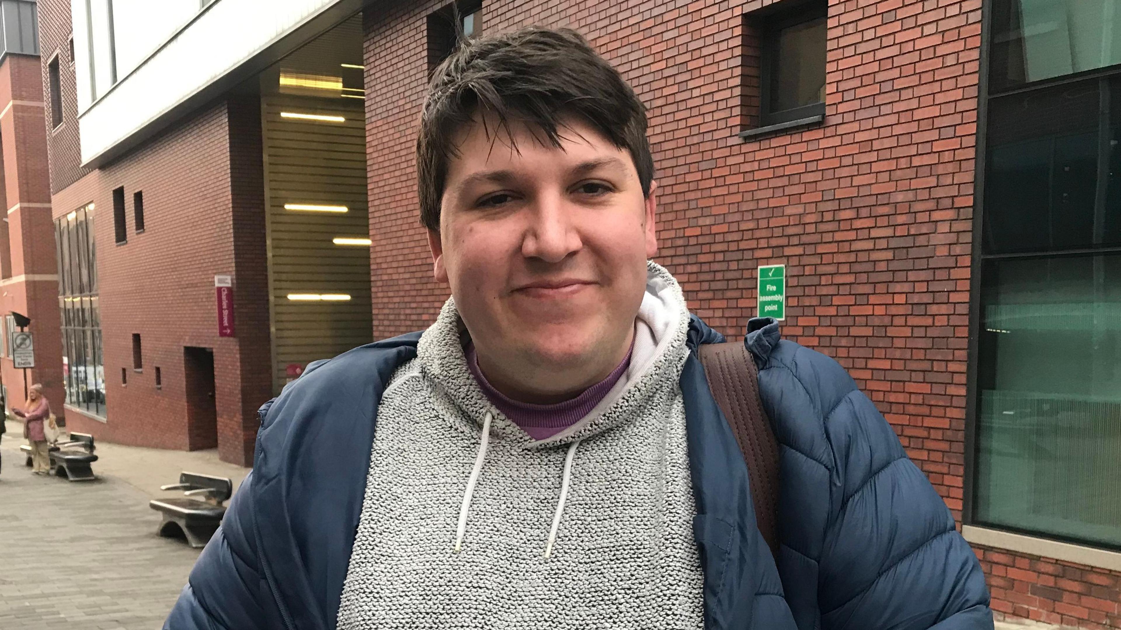 Mr Colburn wears a grey jumper with a blue puffer coat. He stands in front of a red brick building with two benches lined along a footpath in the background.