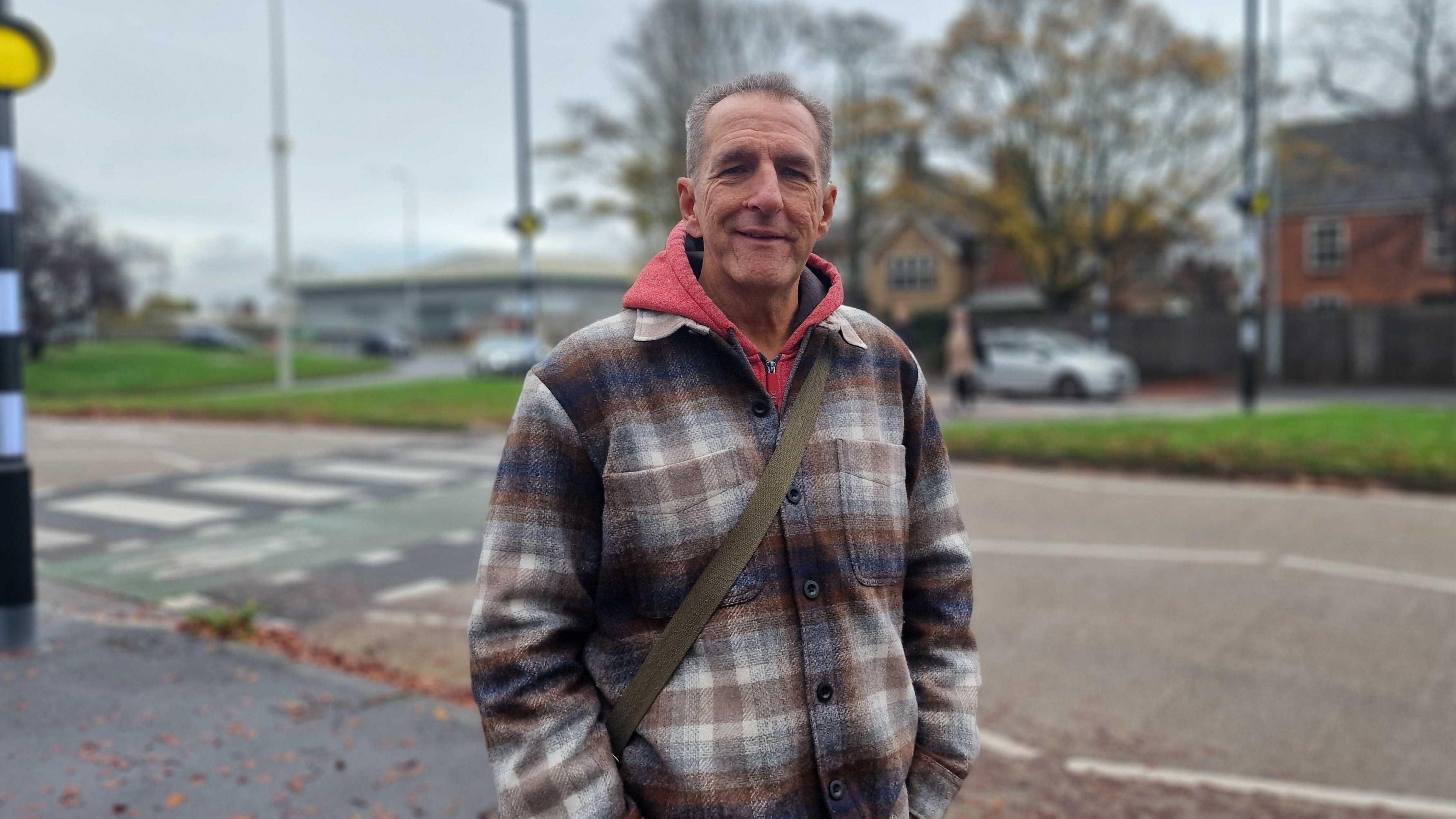 Man with short hair, red hoody, chequered coat and a bag. There is a zebra crossing in the background, a pedestrian, a car, a residential building, a commercial building and trees. There is also a roundabout in the background.