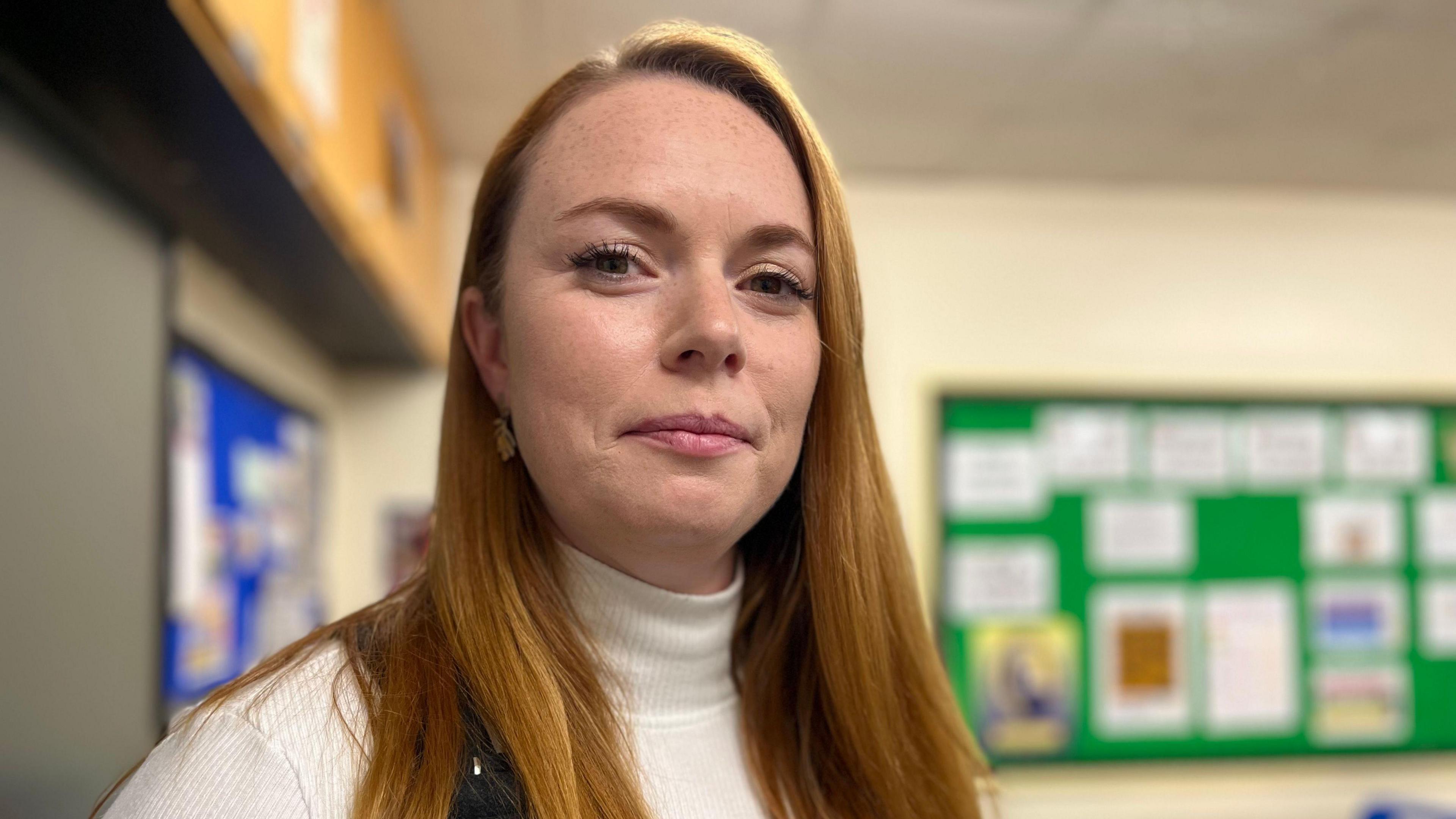 Carla Sheldon is looking at the camera with a closed mouth smile. She has fair skin and long red hair and wears a cream polo neck jumper. She is standing in a classroom, there are blurred display boards behind her.