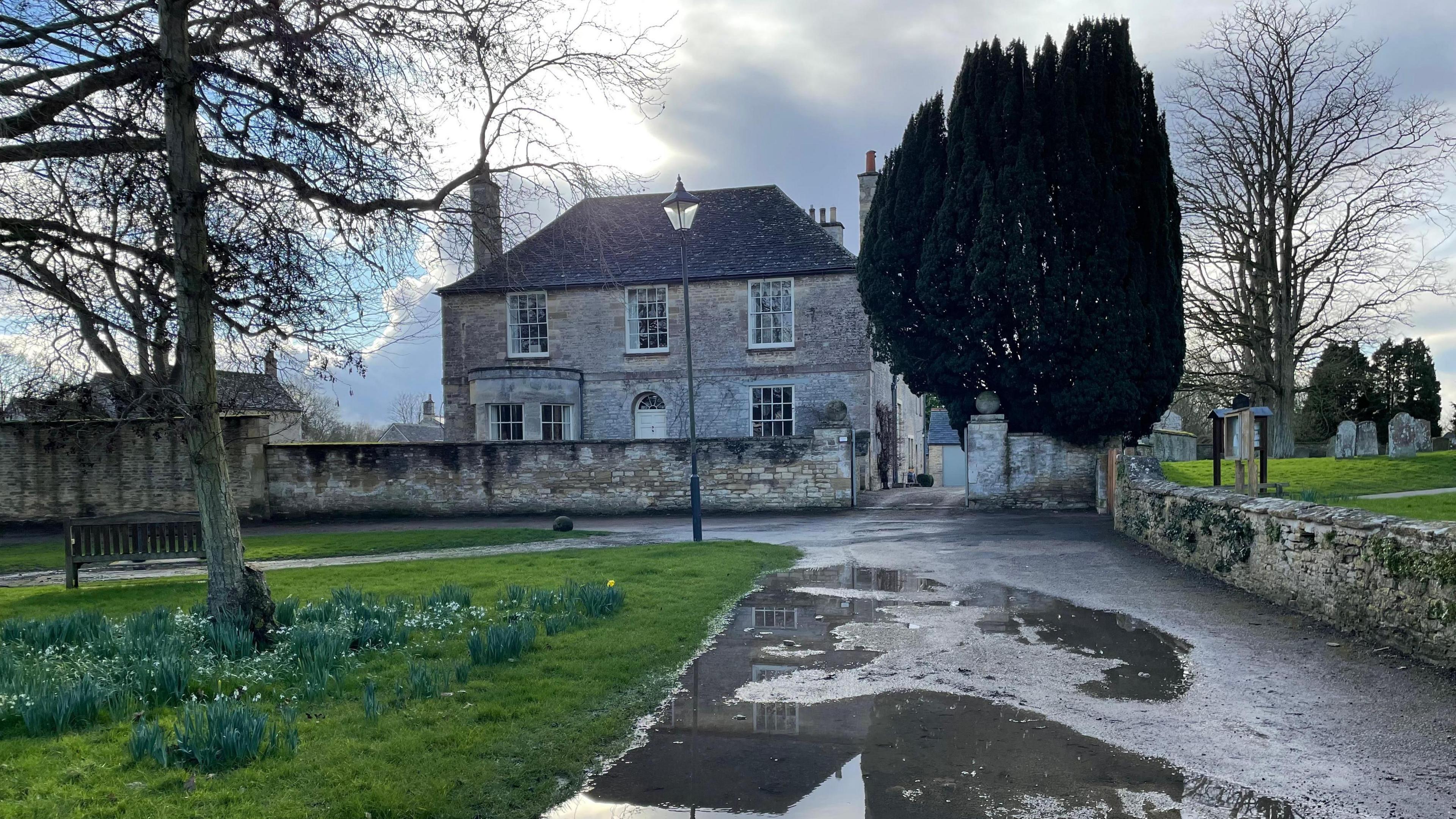 A grey stone house stands behind a low wall. It has two chimneys and the two-storey building has three large upstairs windows. In the foreground is a tree with a road next to it. There is a second tree on a grass verge surrounded by daffodils that are coming into bloom. There are puddles in the road with reflections. Behind the wall beside the house you can see a graveyard.