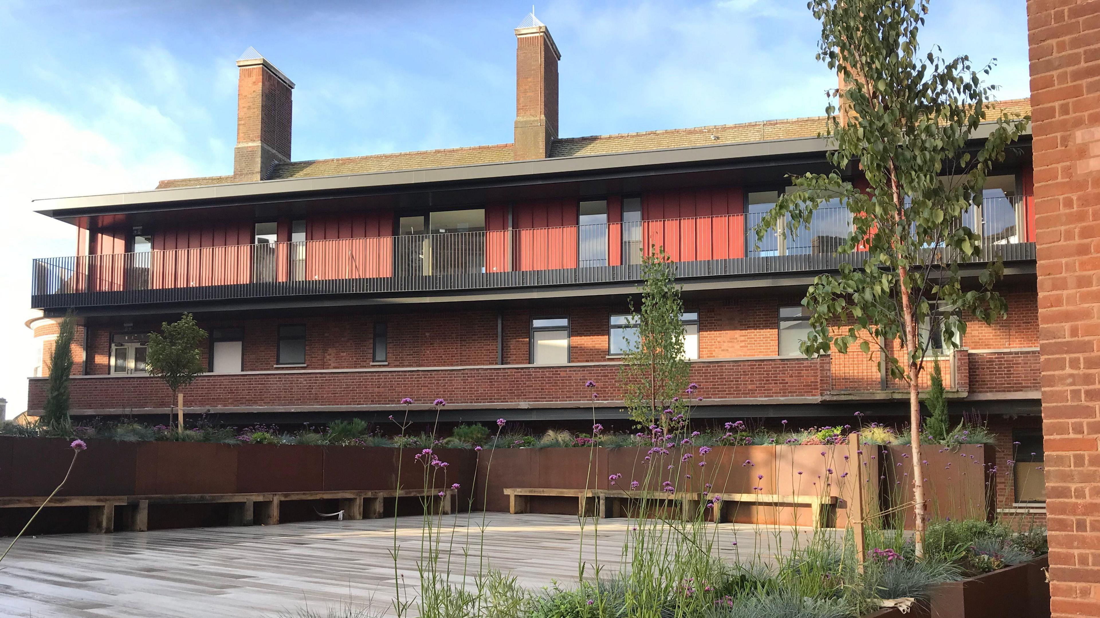 A wooden garden terrace in a luxury apartment block. There are benches along the terrace wall and balconies look onto the garden. Planters also line the perimeter.