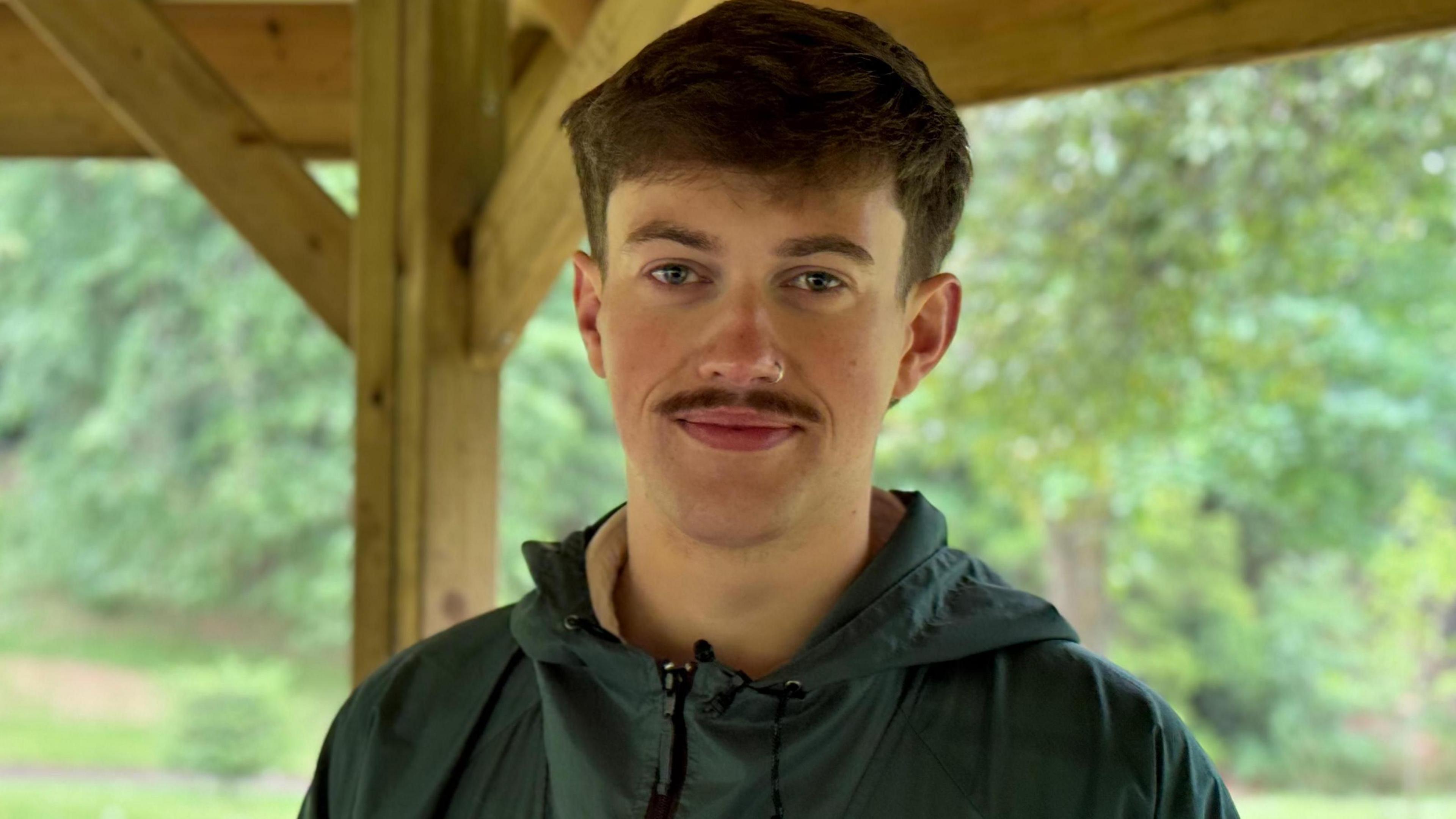 A photo of Lewis looking at the camera. He is outdoors with trees in the background. He has dark hair and a dark thick moustache. He is wearing a green waterproof jacket.