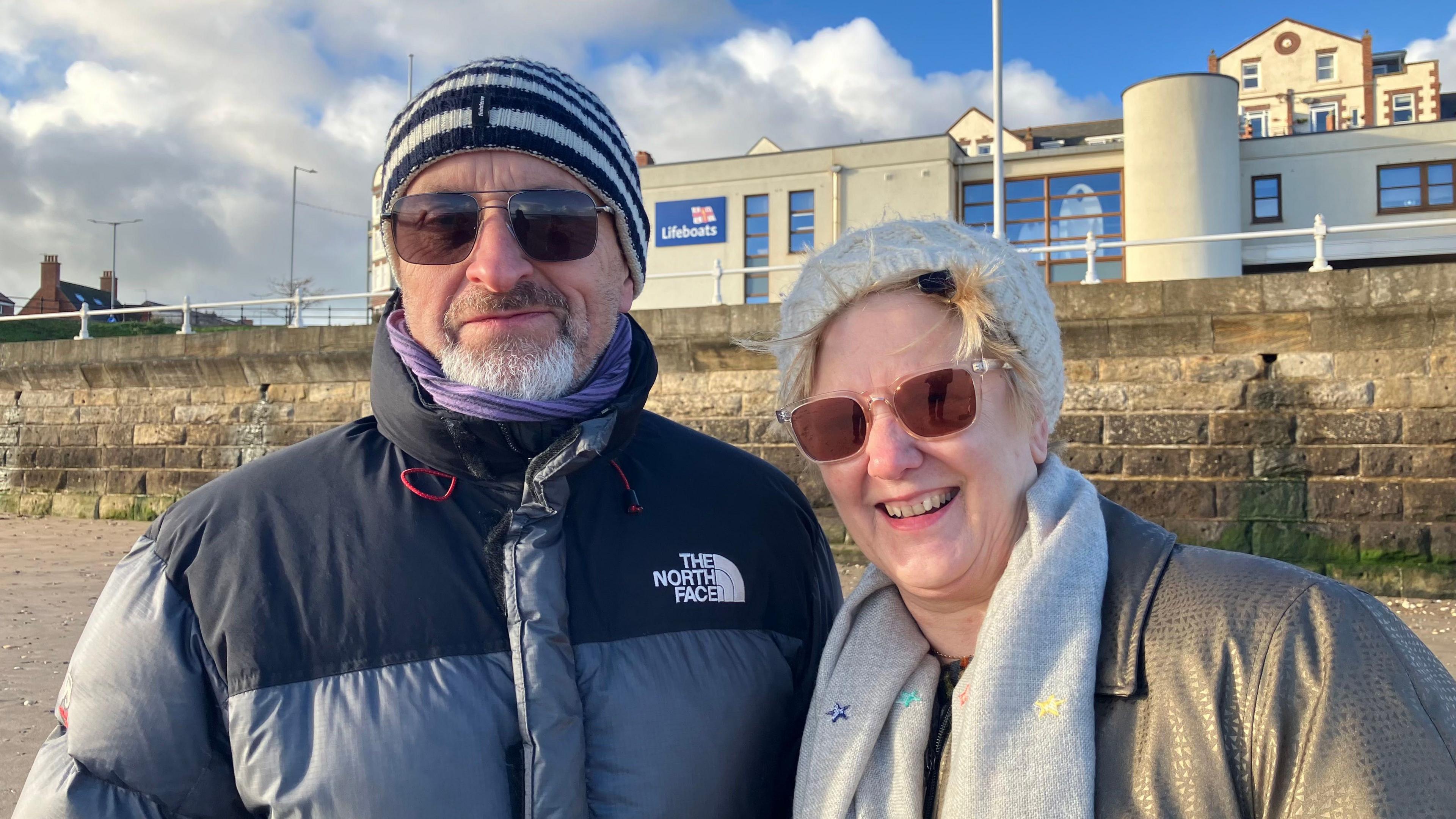 A man with a black and white beanie, short grey beard and brown moustache and sunglasses is wearing a North Face black and grey coat. A woman next to him has a white winter woolly hat on with a grey scarf and khaki coat. You can see a beach wall behind them and a lifeboats building.
