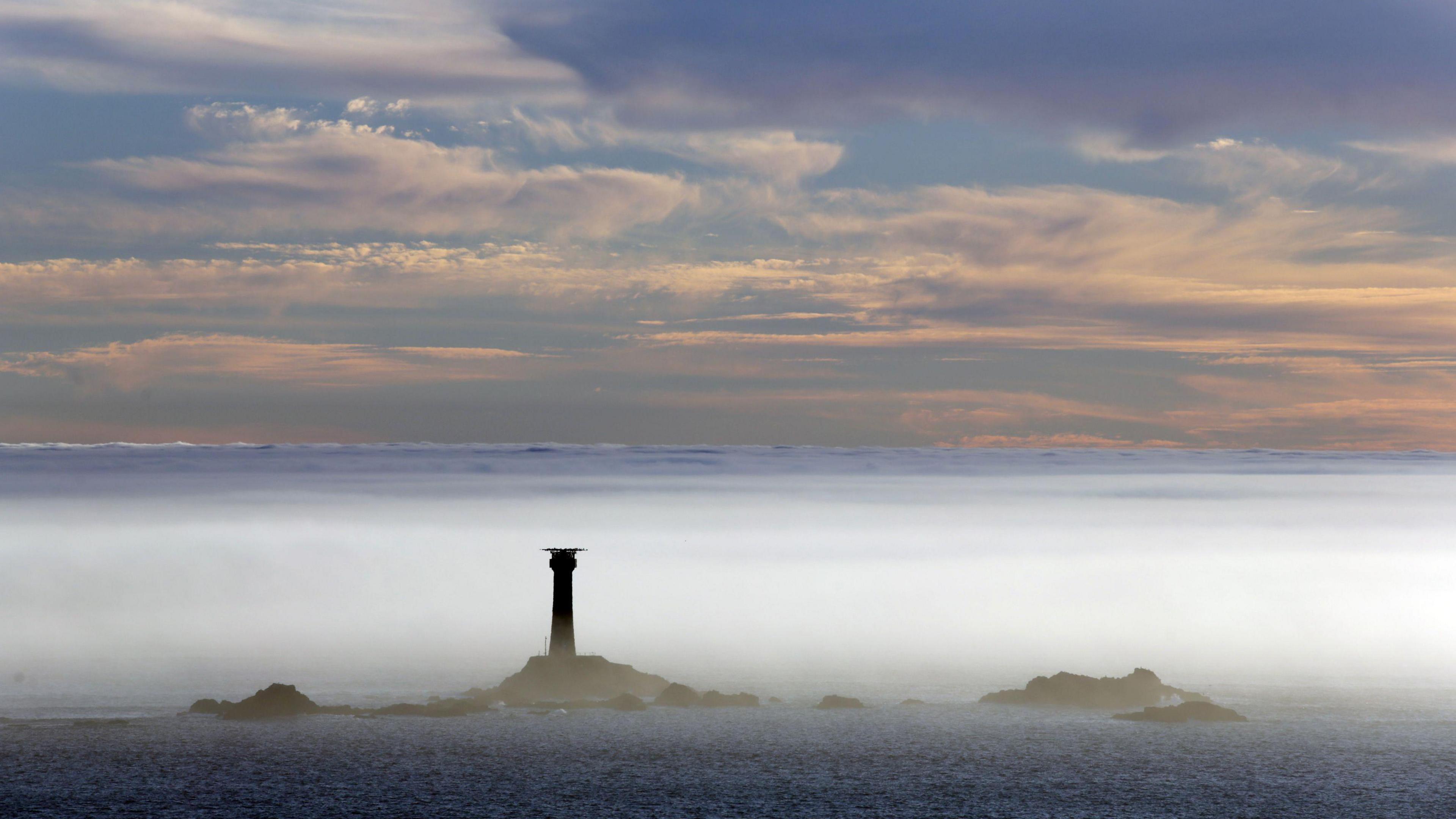 Longships Lighthouse is shrouded in mist and surrounded by a calm sea. Above the layer of fog is a pale blue sky filled with pale link clouds.