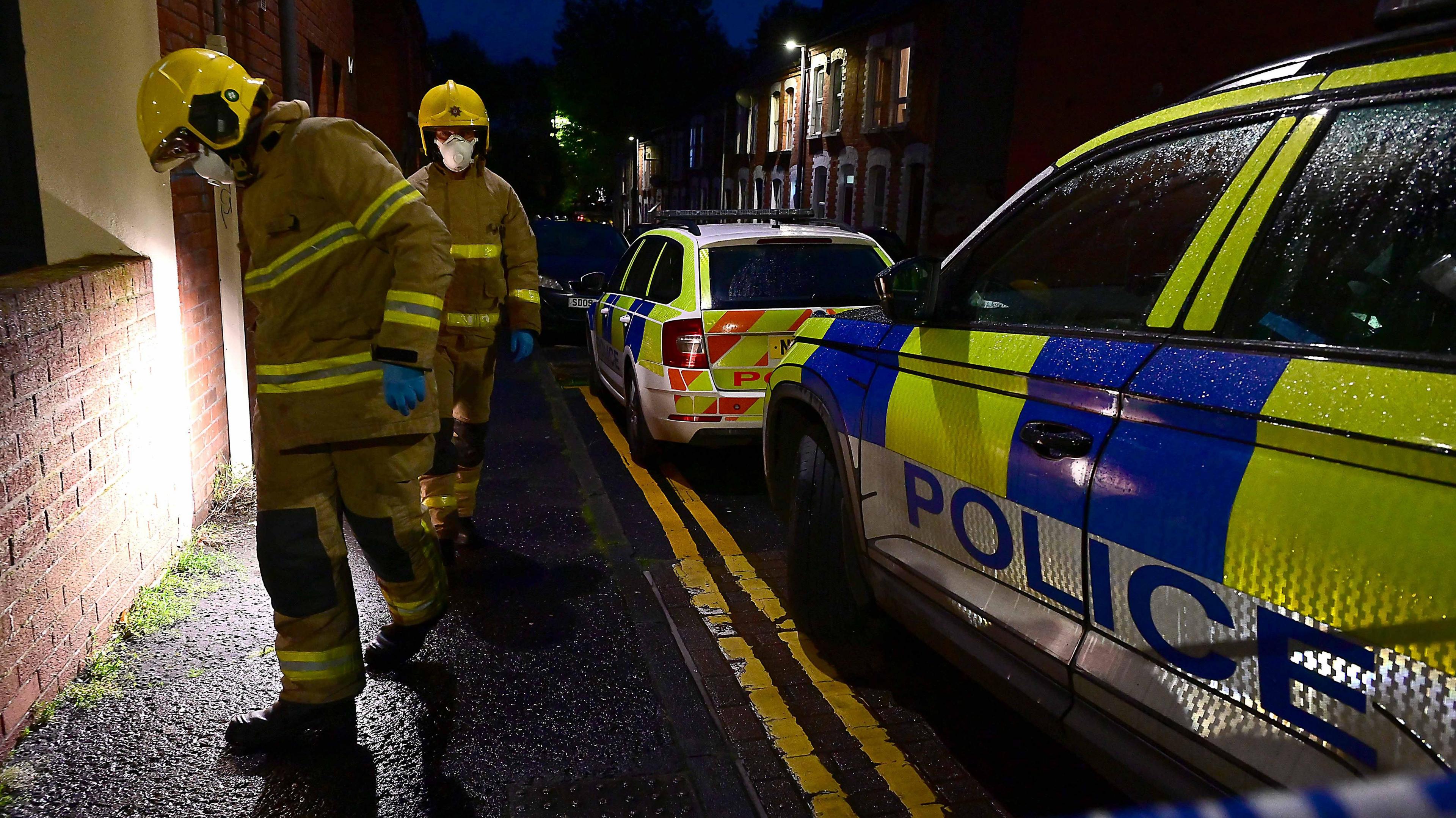 Two firefighters wearing face masks walk alongside two marked police cars as a light is shone on a brick wall on Damascus Street.