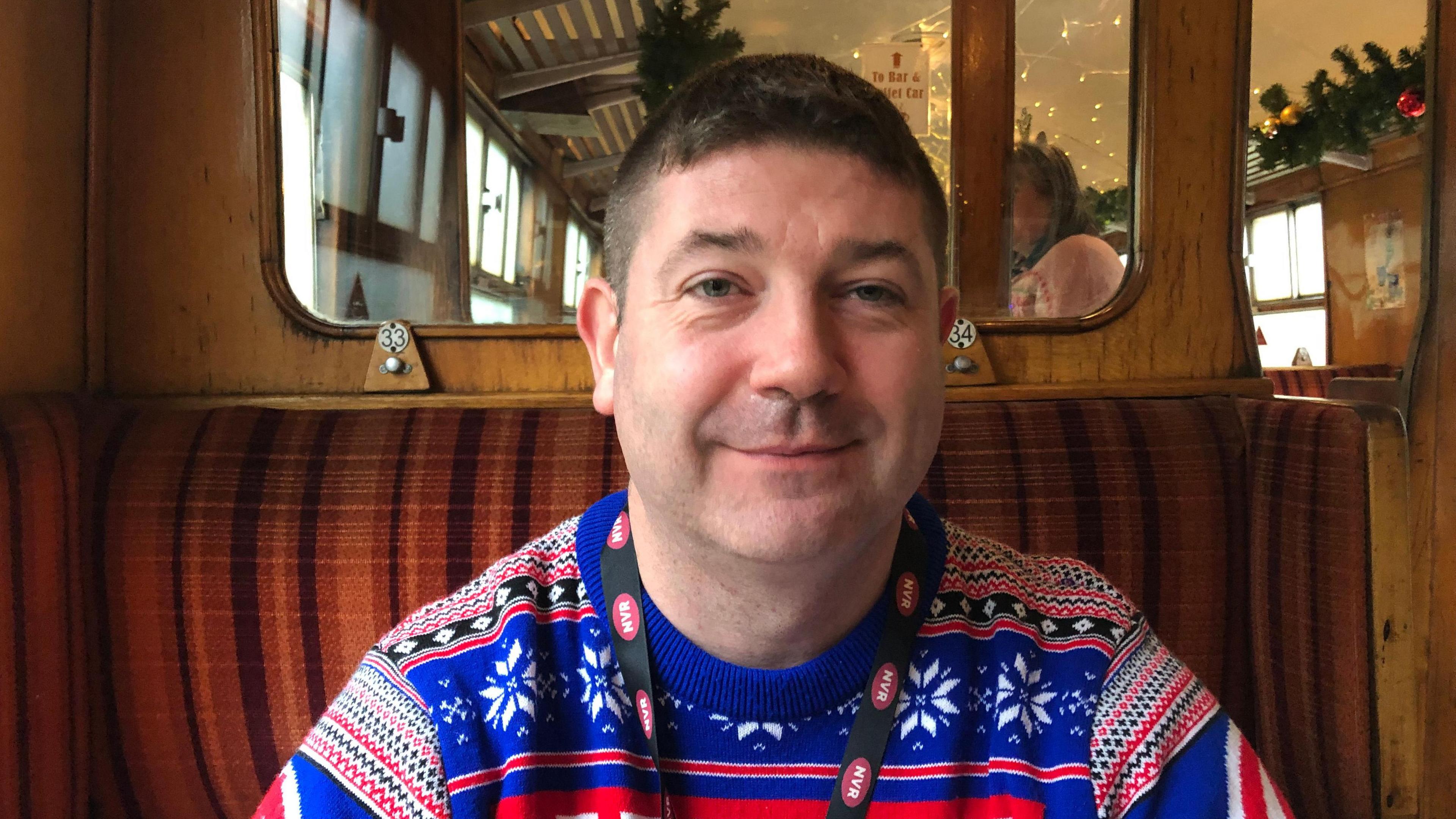 Chris O'Regan has short black hair and is wearing a blue, red and white Christmas jumper, with an icicle pattern on it. He has an NVR lanyard around his neck and is sitting in an old railway carriage