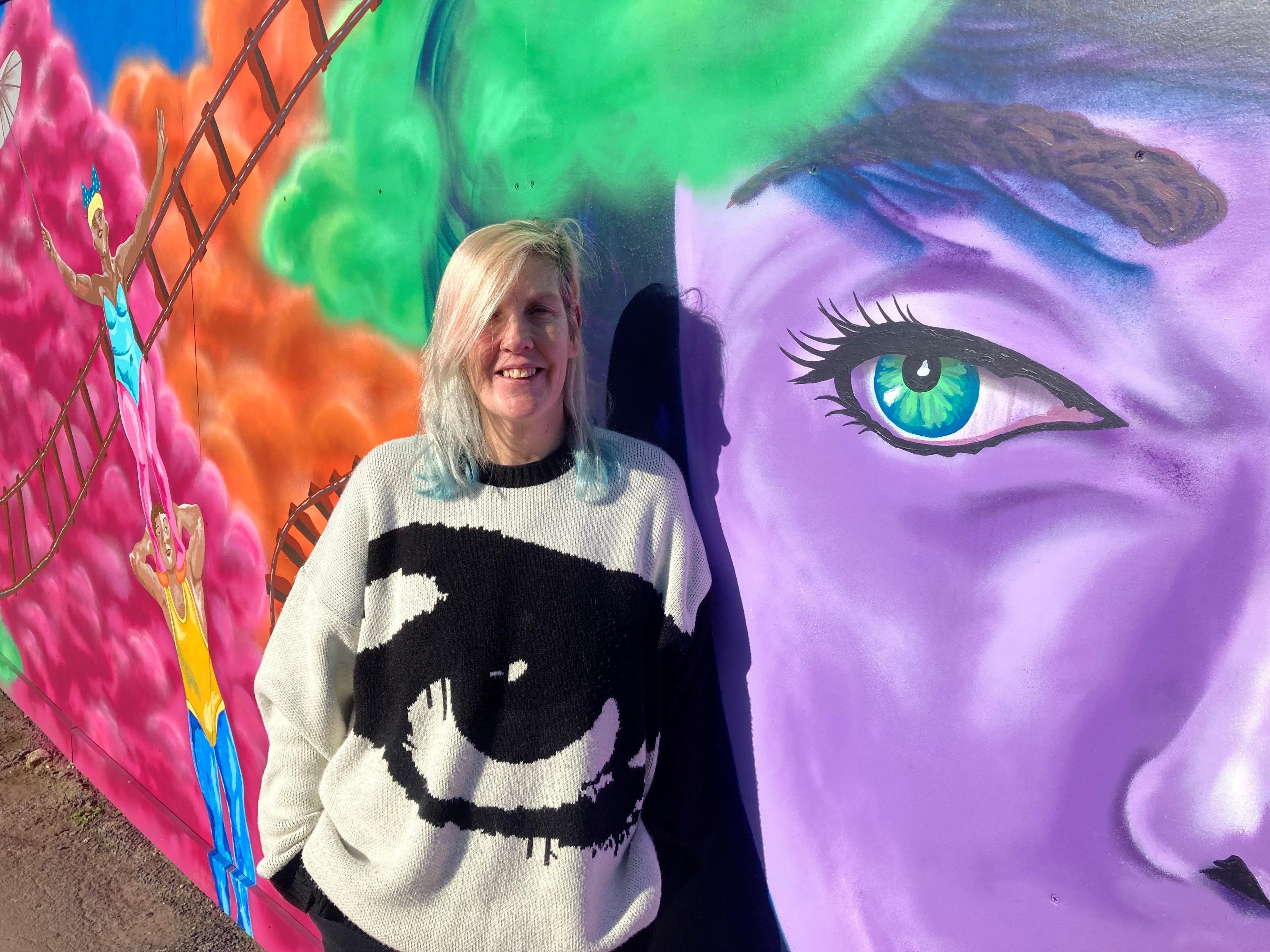 Street artist, Sarah Harris, stood against a wall displaying her artwork in Swindon. The painting includes a huge purple face with green eyes, a train track cutting through colourful clouds and the well known Swindon blondinie statue