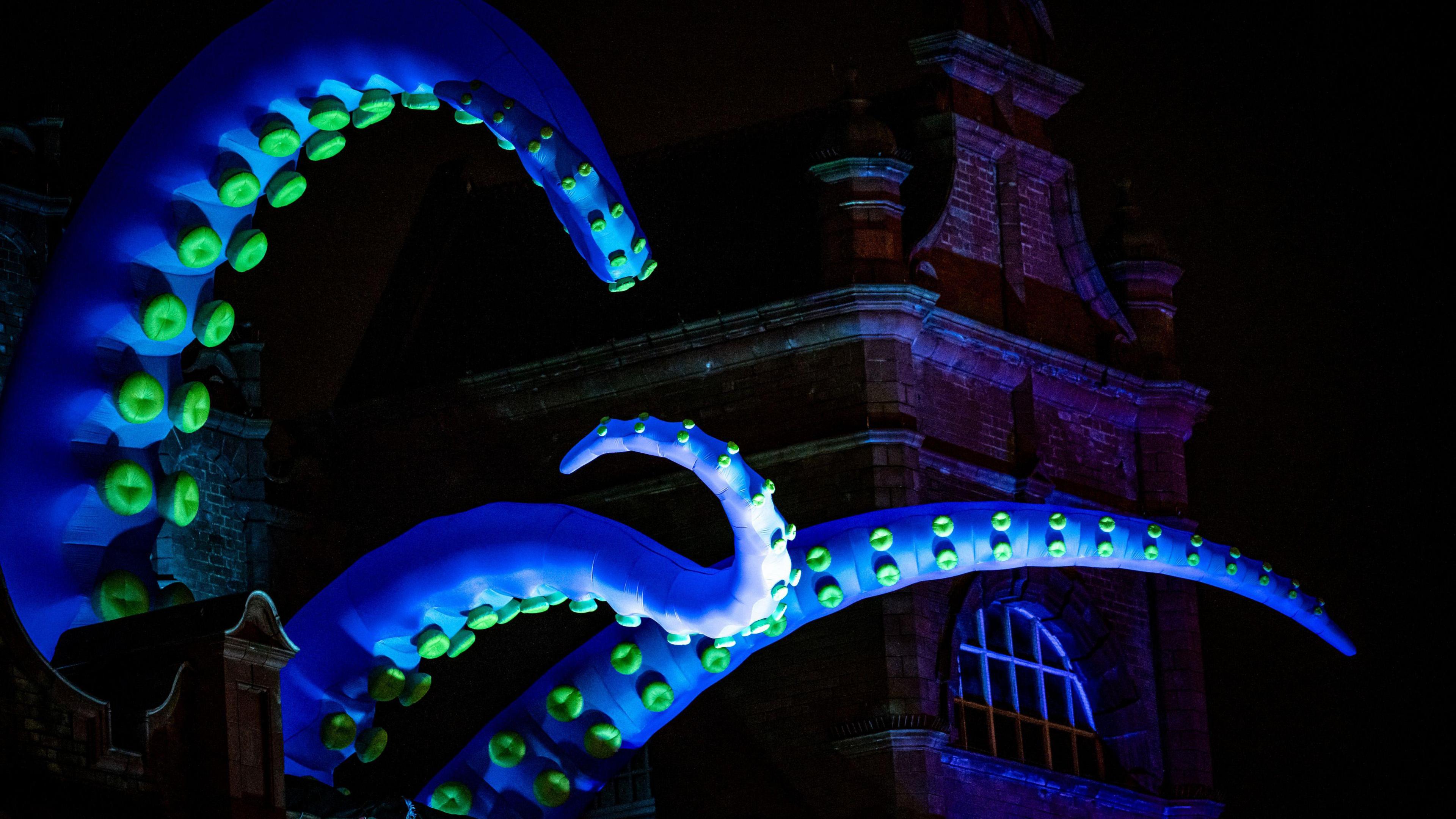 Blue inflatable tentacles from a giant inflatable creature attached to a tall building in Morecambe at night.