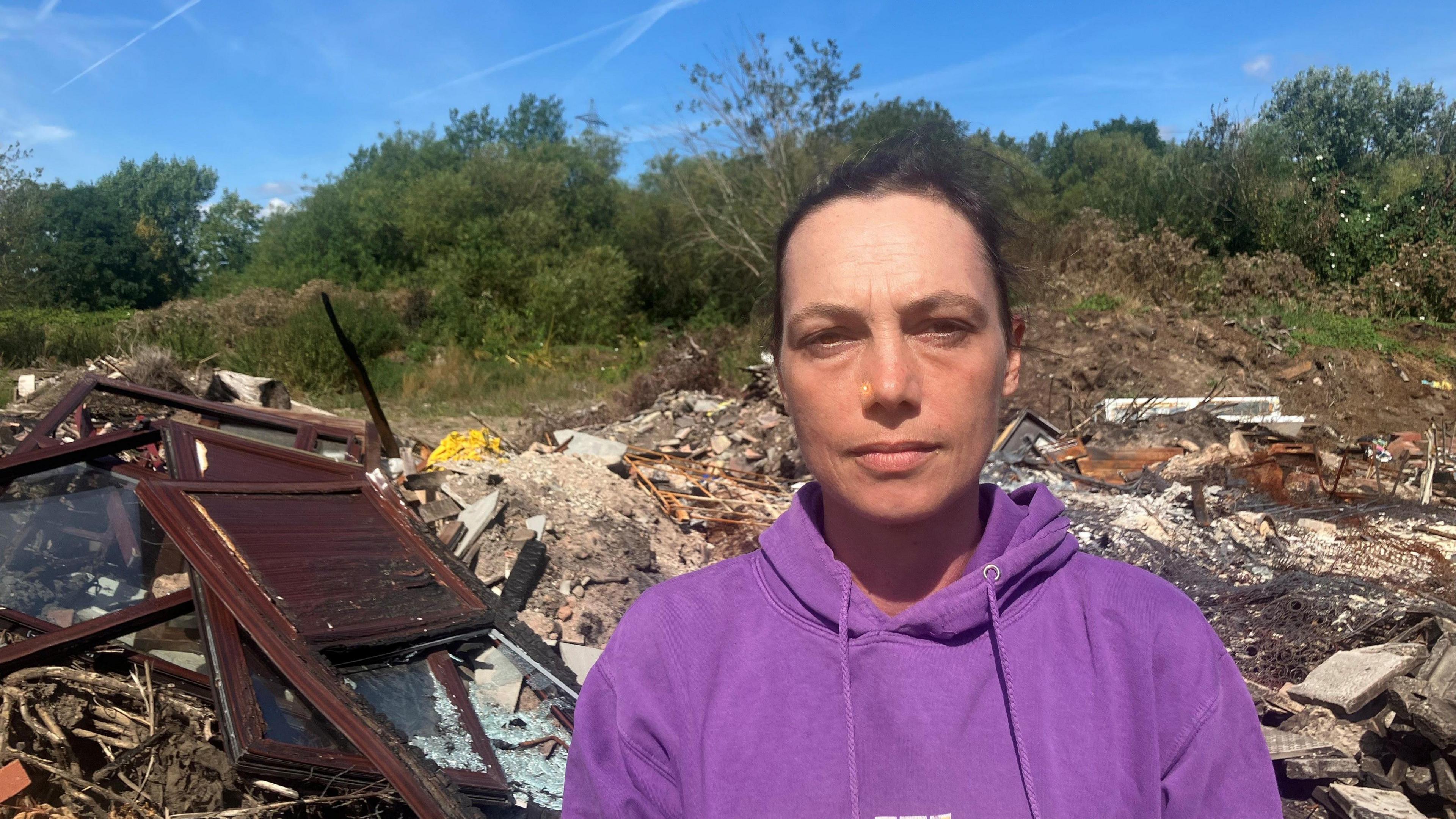 Photograph of Helen Carr from Flixton. She is pictured in front of a suspected illegal waste dump close to the River Mersey. She has tied-back dark hair and is wearing a purple hooded top.