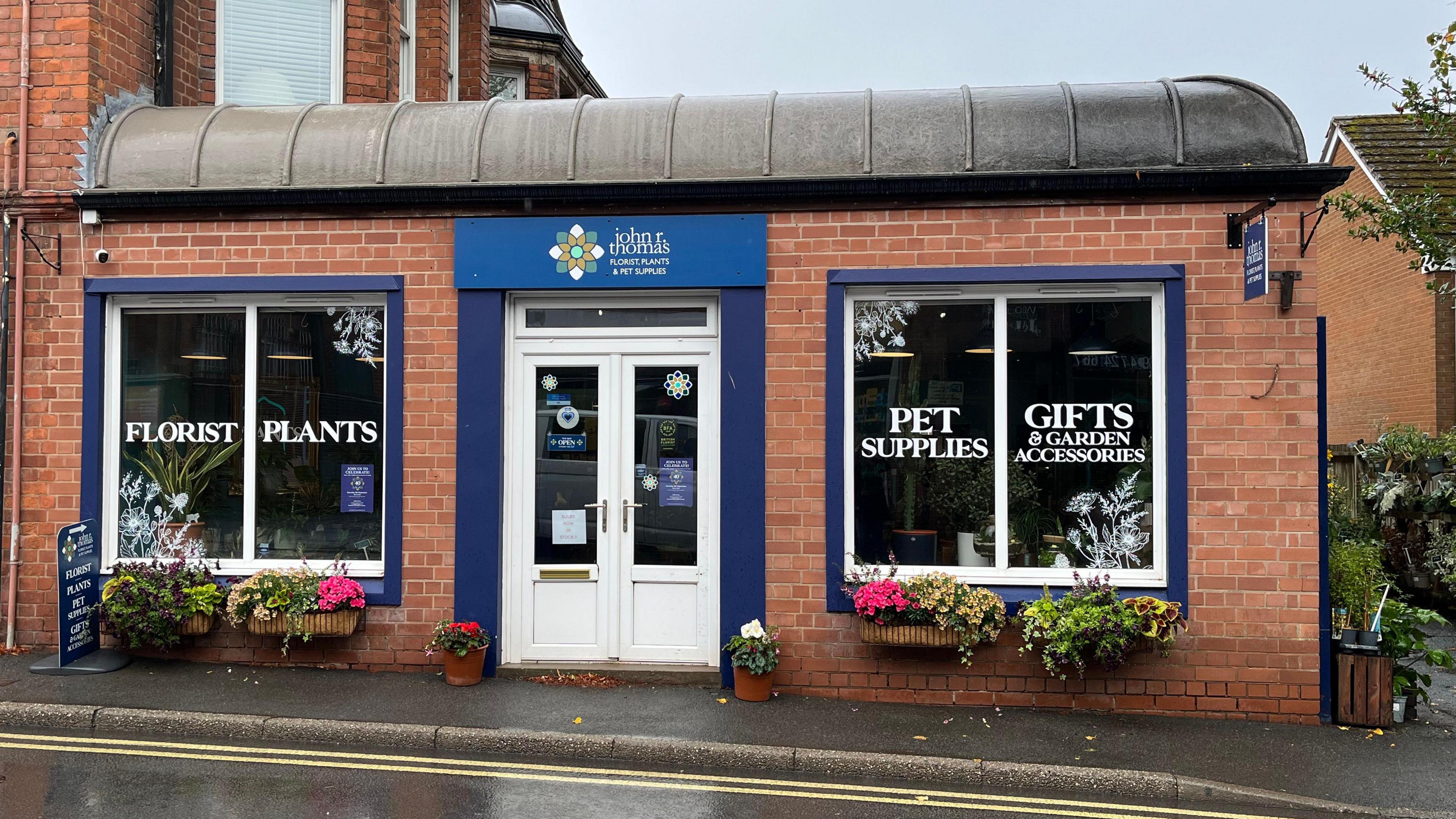 A single-storey red brick building with dark blue frames around the windows and door. The door is white, with a blue sign above it reading 'John R Thomas' in white lettering. On the four windows, the words 'florist', 'plants', 'pet supplies', and 'gifts & garden accessories' are printed. There are four window boxes outside with bright pink and yellow flowers, and two pot plants sit by the door. The sky is grey, overcast, and the wet pavement indicates it's recently been raining.