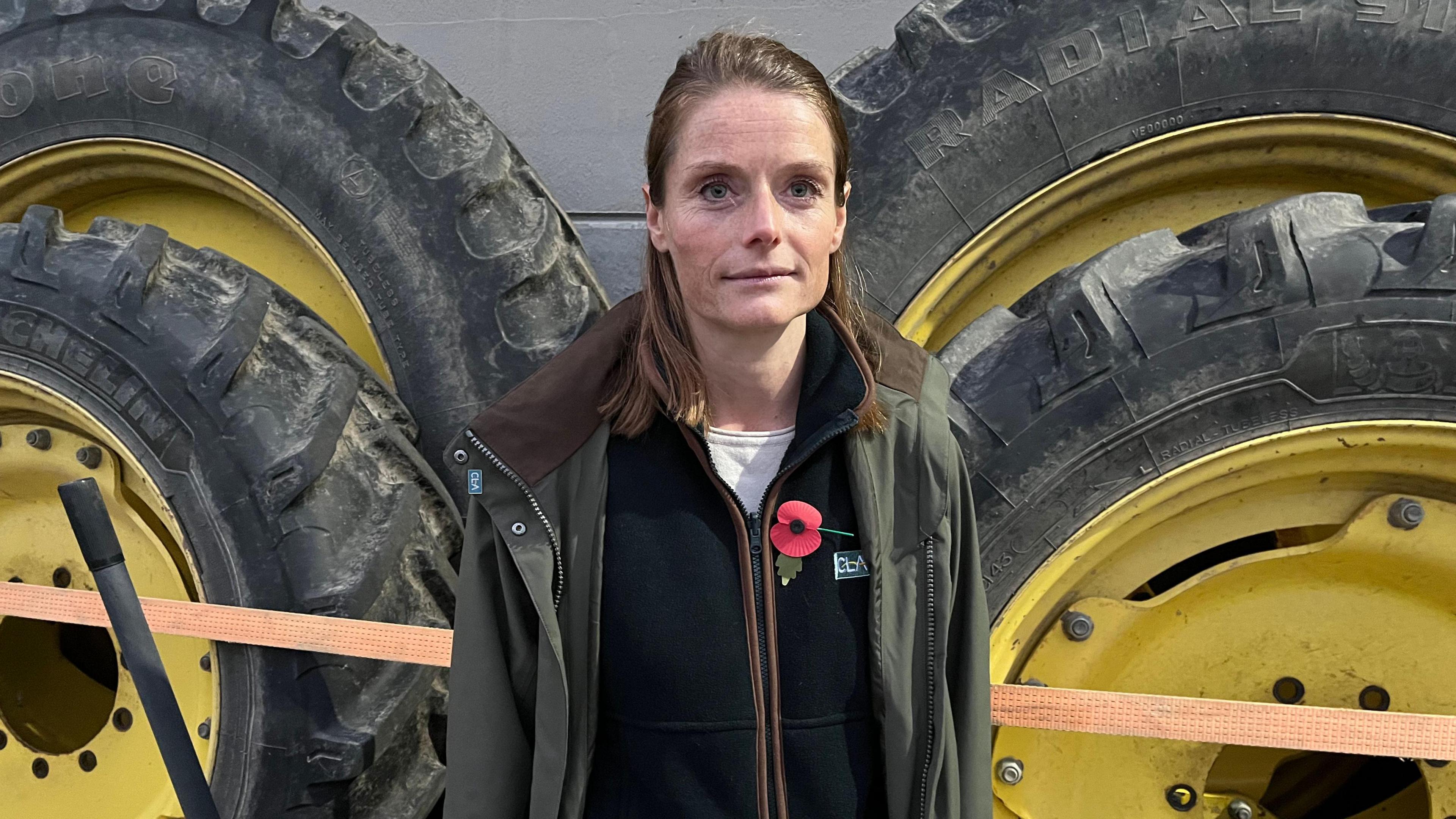 Cath Crowther looks to the right of the camera, she is wearing a green overcoat, black fleece, white top and with a poppy on her left side. She is standing in front of four tractor's wheels which are propped up against a wall. She has brown hair and blue eyes. 