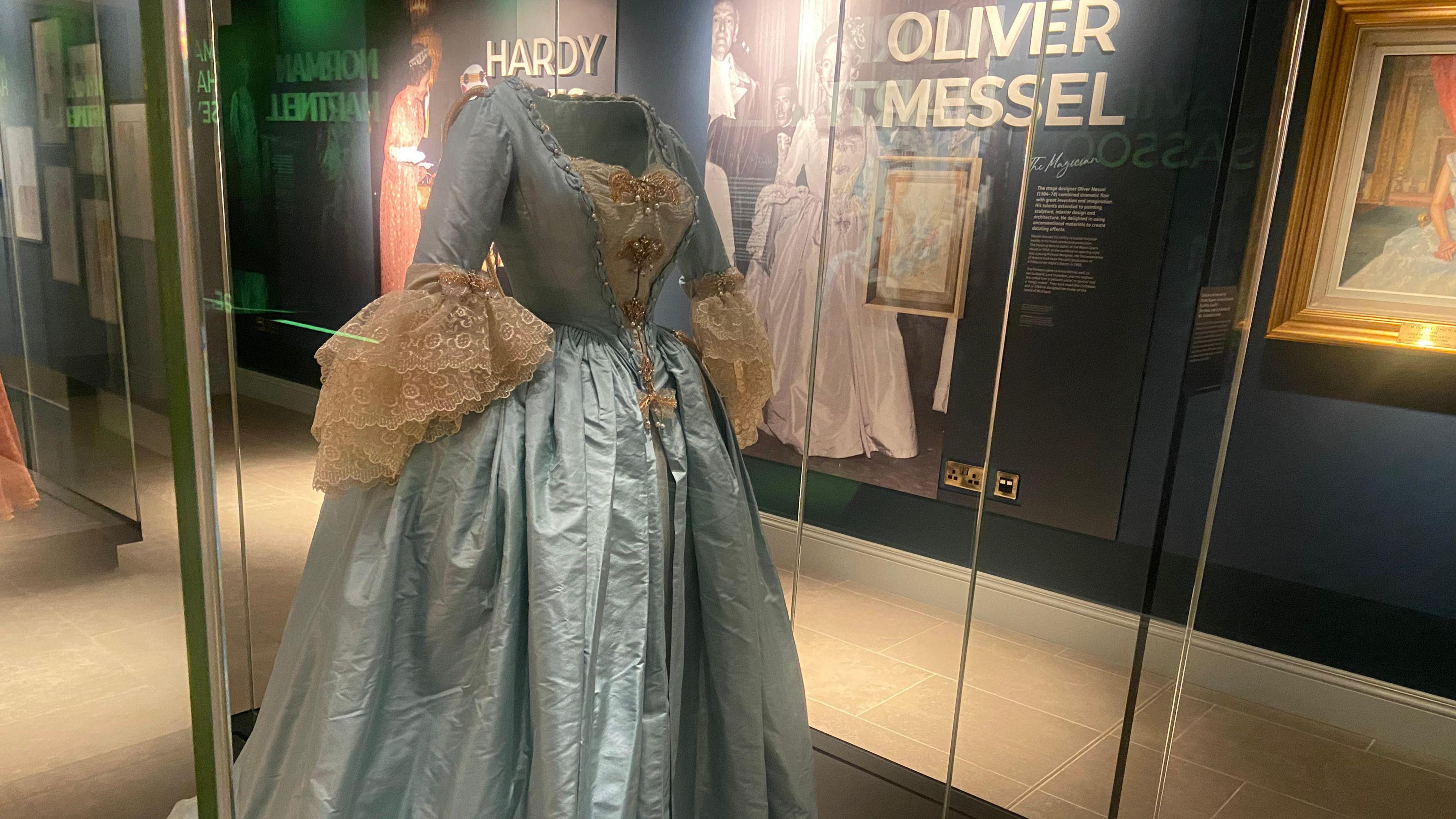 An eighteenth-century-style blue and gold ballgown designed by Oliver Messel and worn by Princess Margaret to a charity ball at London’s Mansion House in July 1964. An exhibition sign for Oliver Messel with a picture of Princess Margaret wearing the gown can be seen in the background.