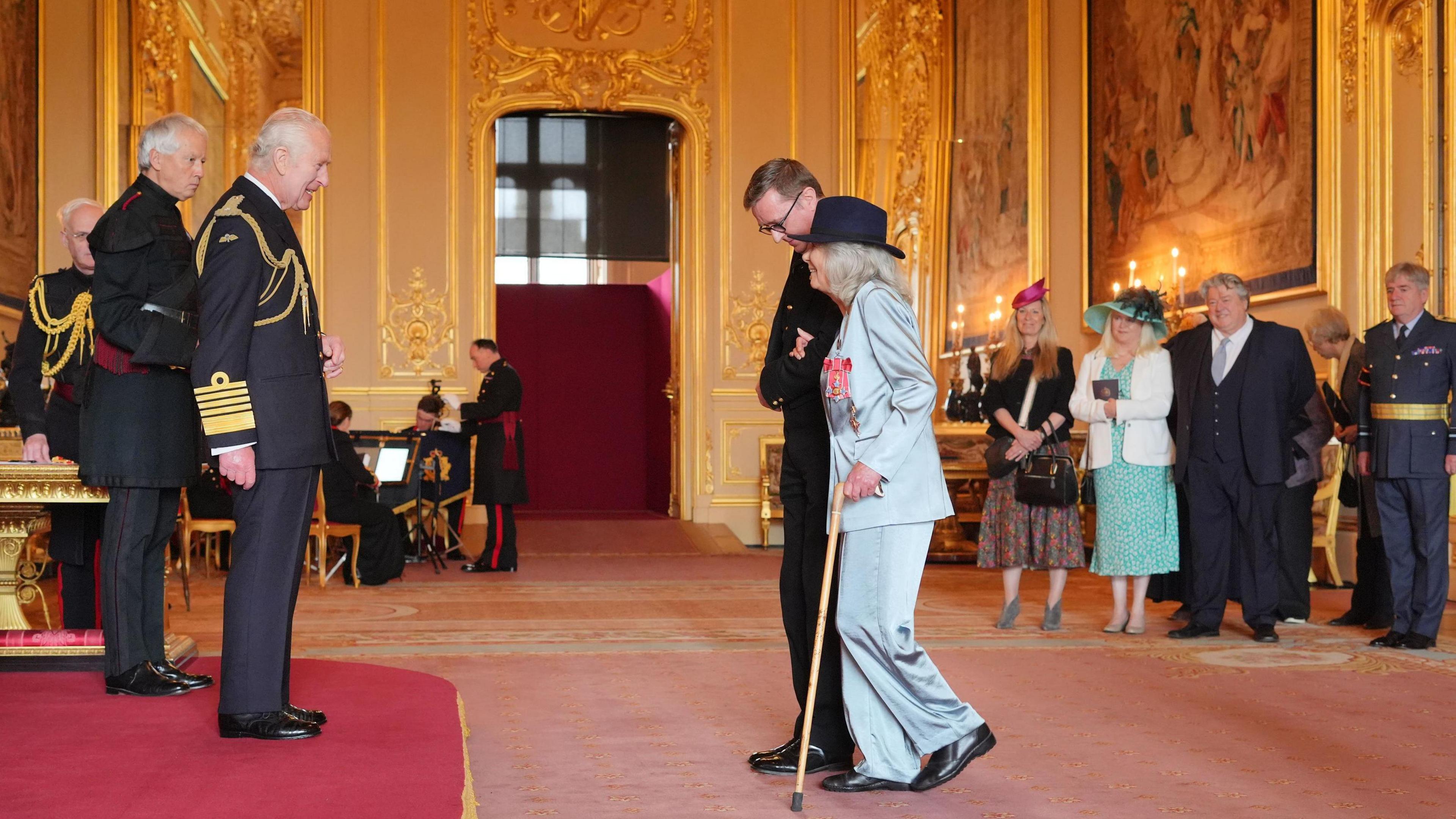 Dame Jilly Cooper, from Stroud, Author, is made a Dame Commander of the British Empire by King Charles III at Windsor Castle. She is walking towards the King, using a stick.