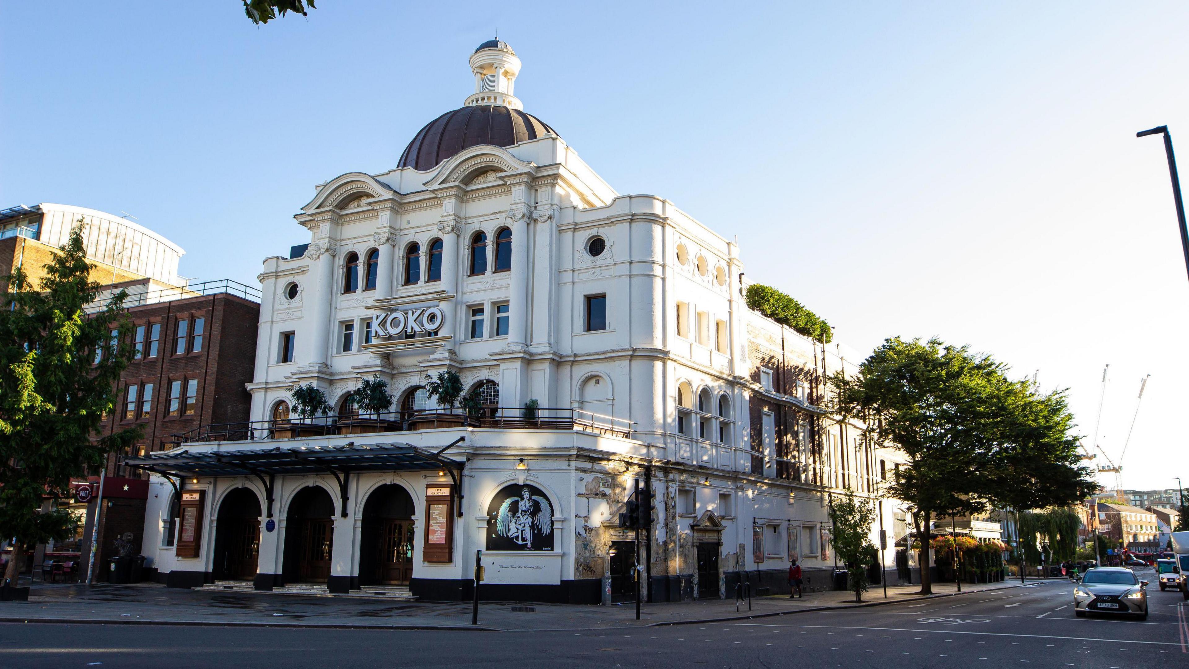 Wide shot of the outside of Koko music venue, a white building formerly known as the Camden Palace that sits on the corner of the road. The facade is in a Baroque pastiche style with three arched entrances and an awning above them.