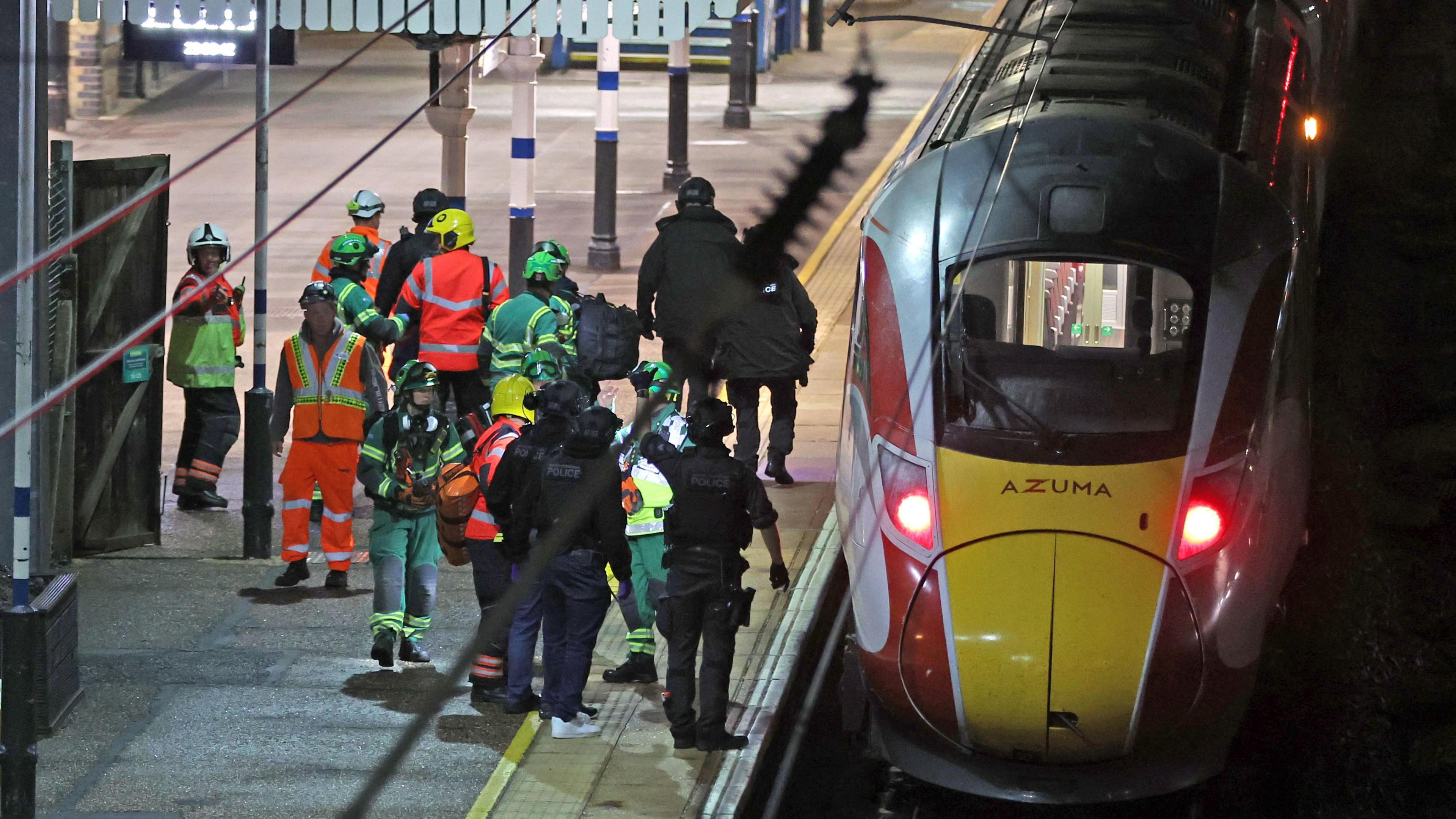 A group of people in high-vis green and orange vests and jackets crowd on a platform beside a train. It is after dark.