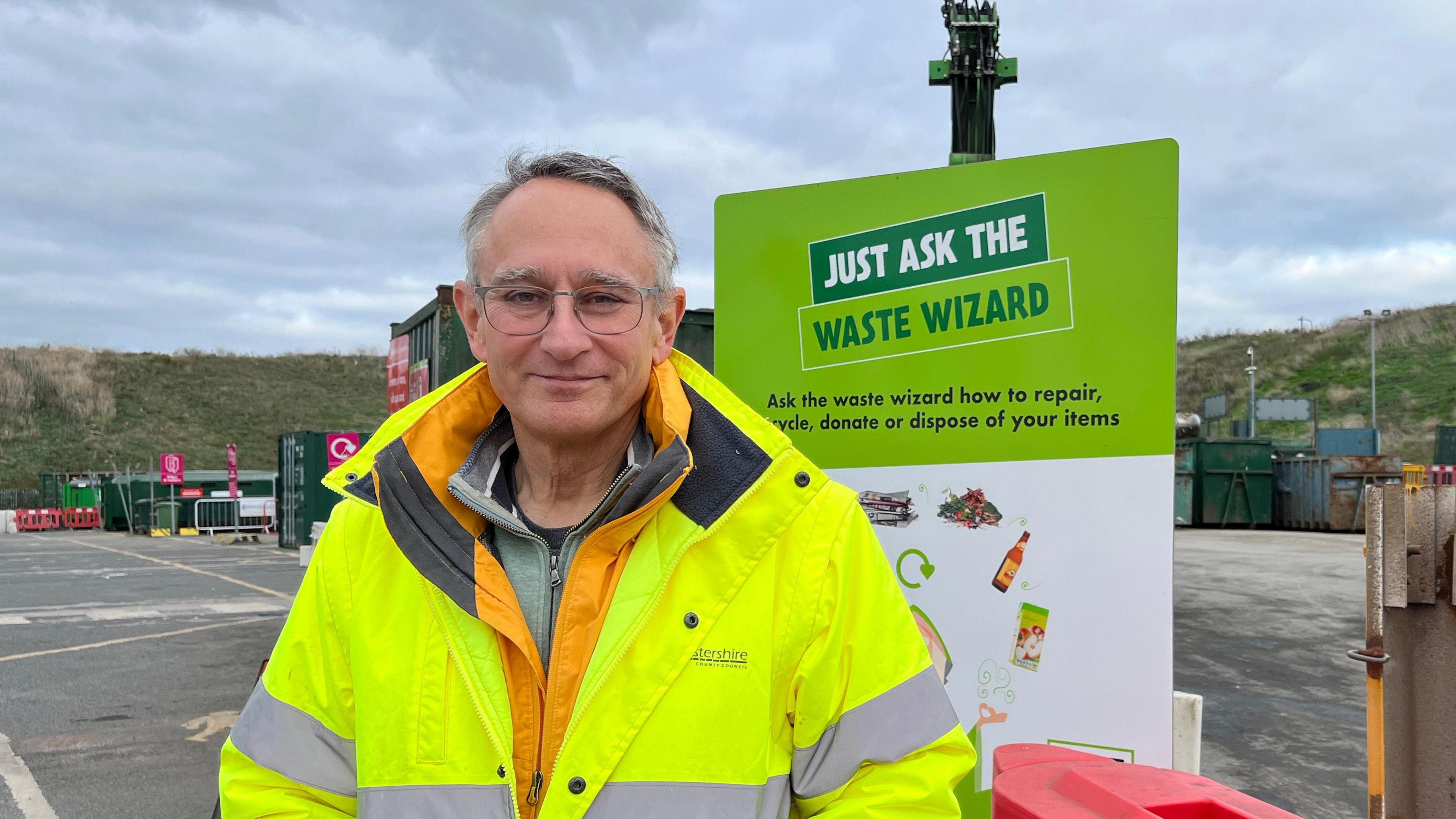 Councillor Martin Horwood stood at the recycling centre. He is wearing a hi-vis jacket and is looking directly into the camera.