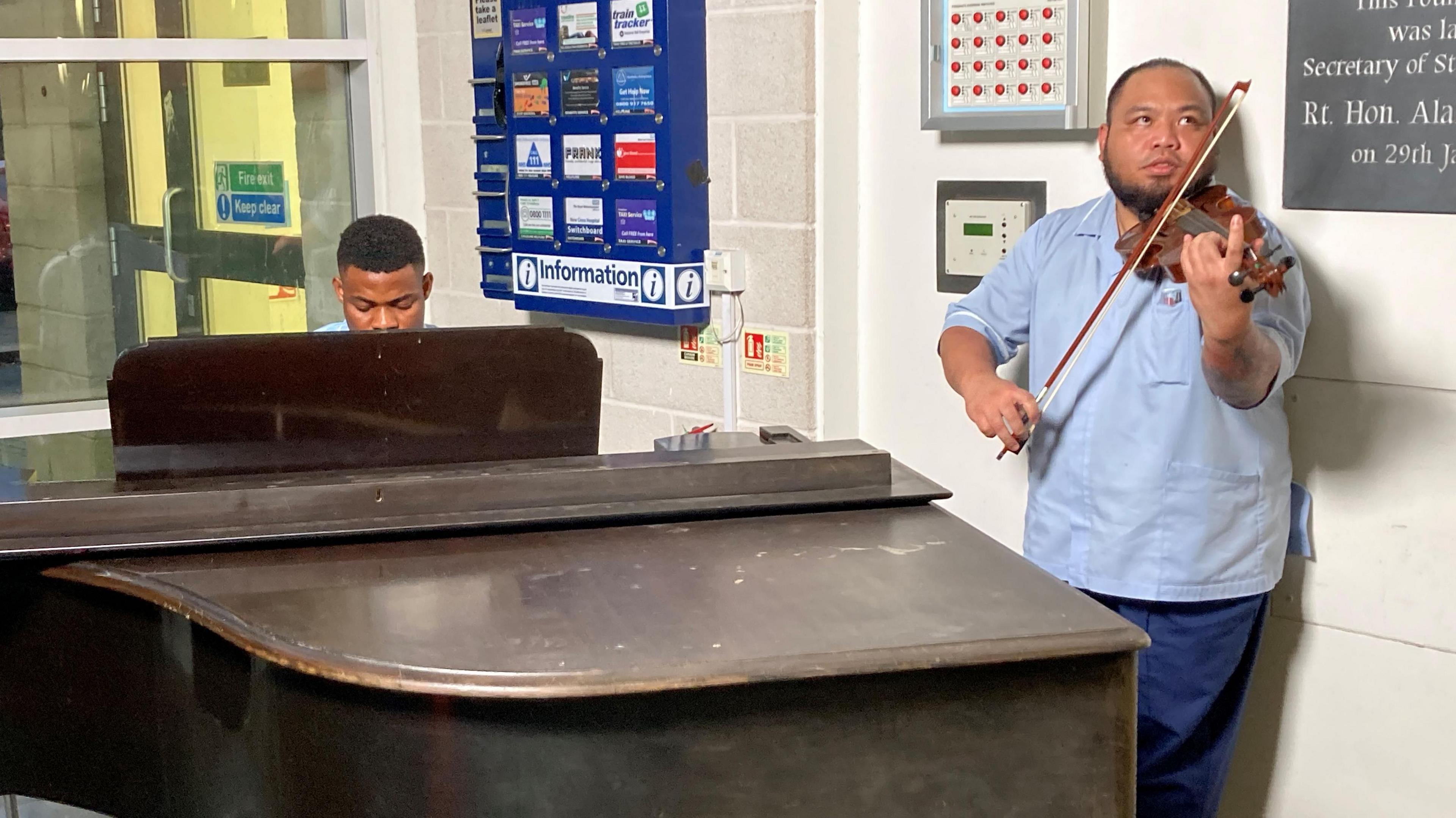 A man sits at a baby grand piano and another man plays the violin. Both wear blue nurses' uniforms. 