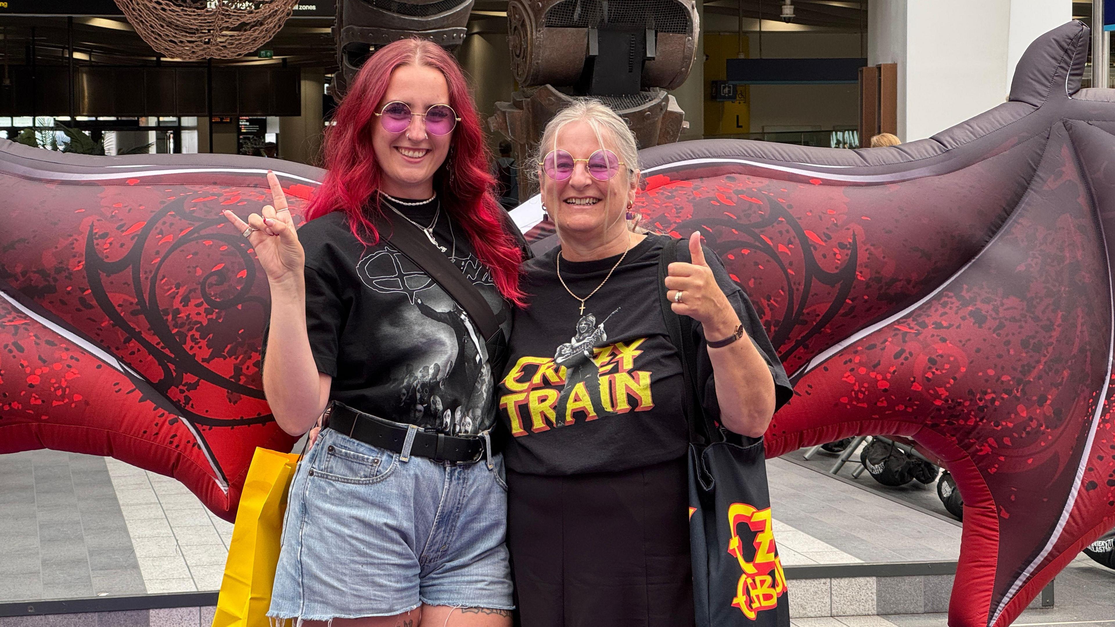 A woman with red hair stands next to an older woman with blonde hair. They both grin at the camera, wearing black Ozzy Osbourne T-shirts and round pink glasses. They stand in front of an inflatable pair of red wings.