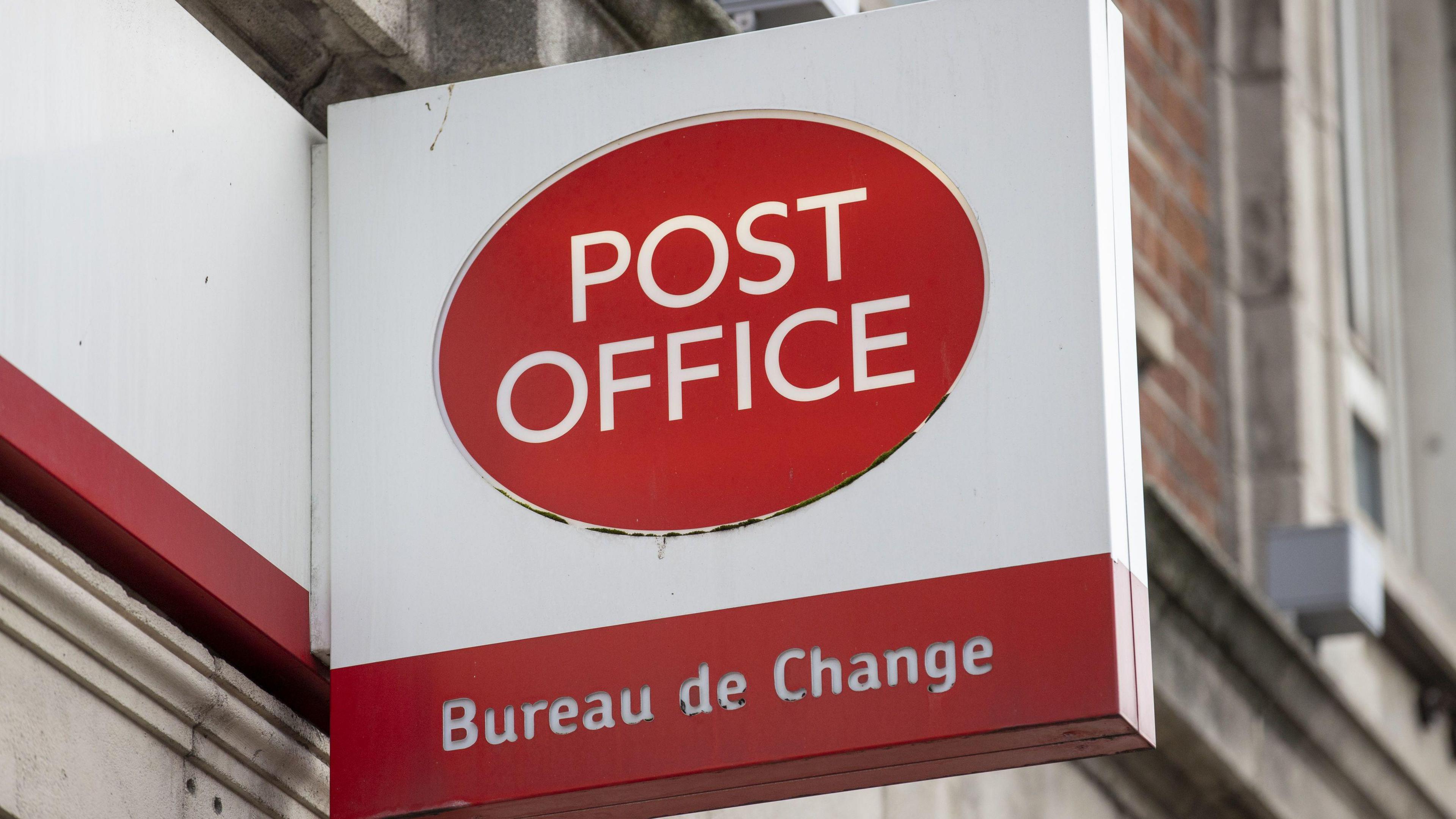 A red circle on a white sign. In the red circle are the words 'post office' in white. Behind the sign are brick walls.