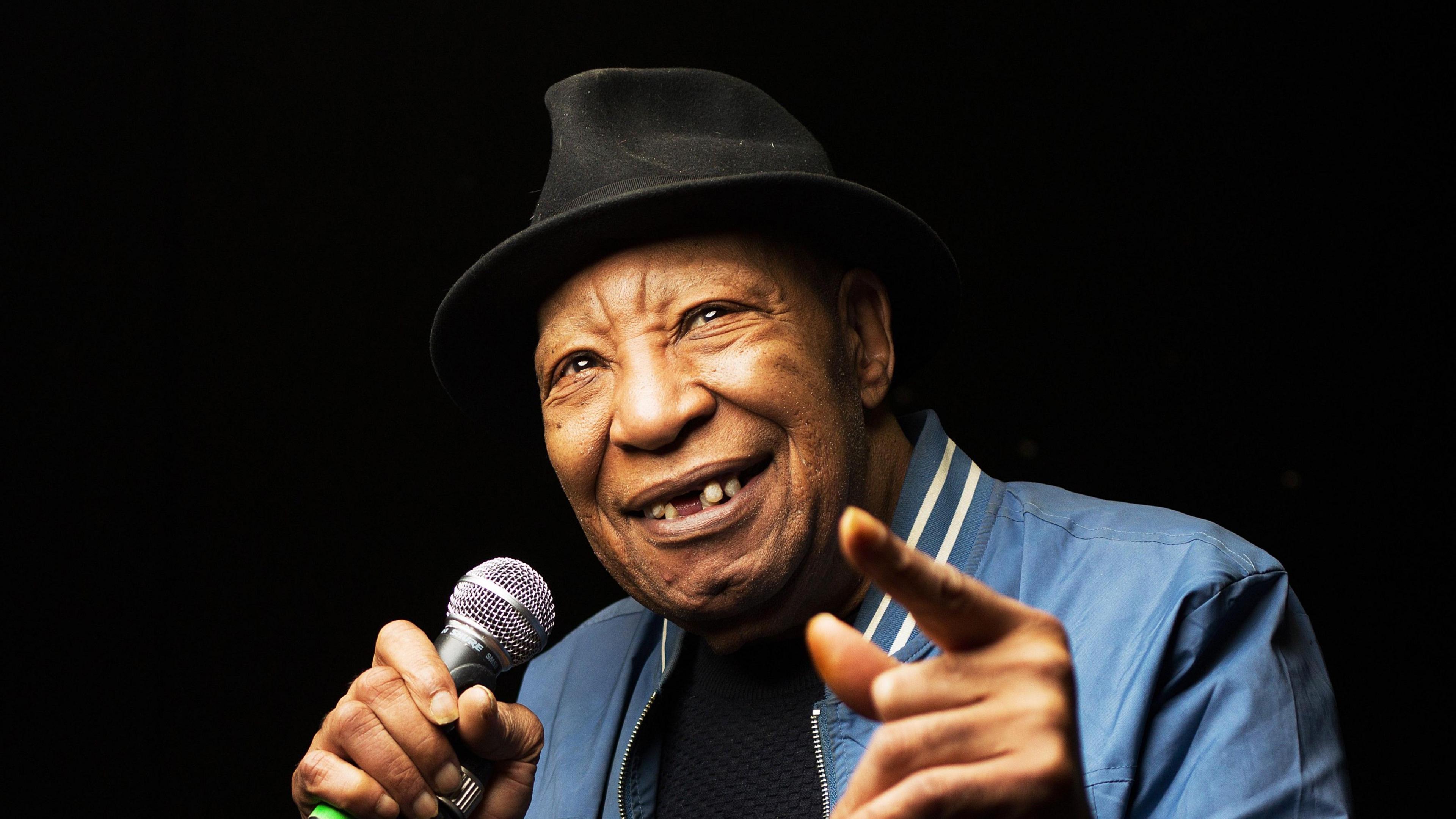 Geno Washington smiles as he holds a microphone in one hand and points a finger in front of him. He wears a black hat, blue jacket and navy jumper underneath.