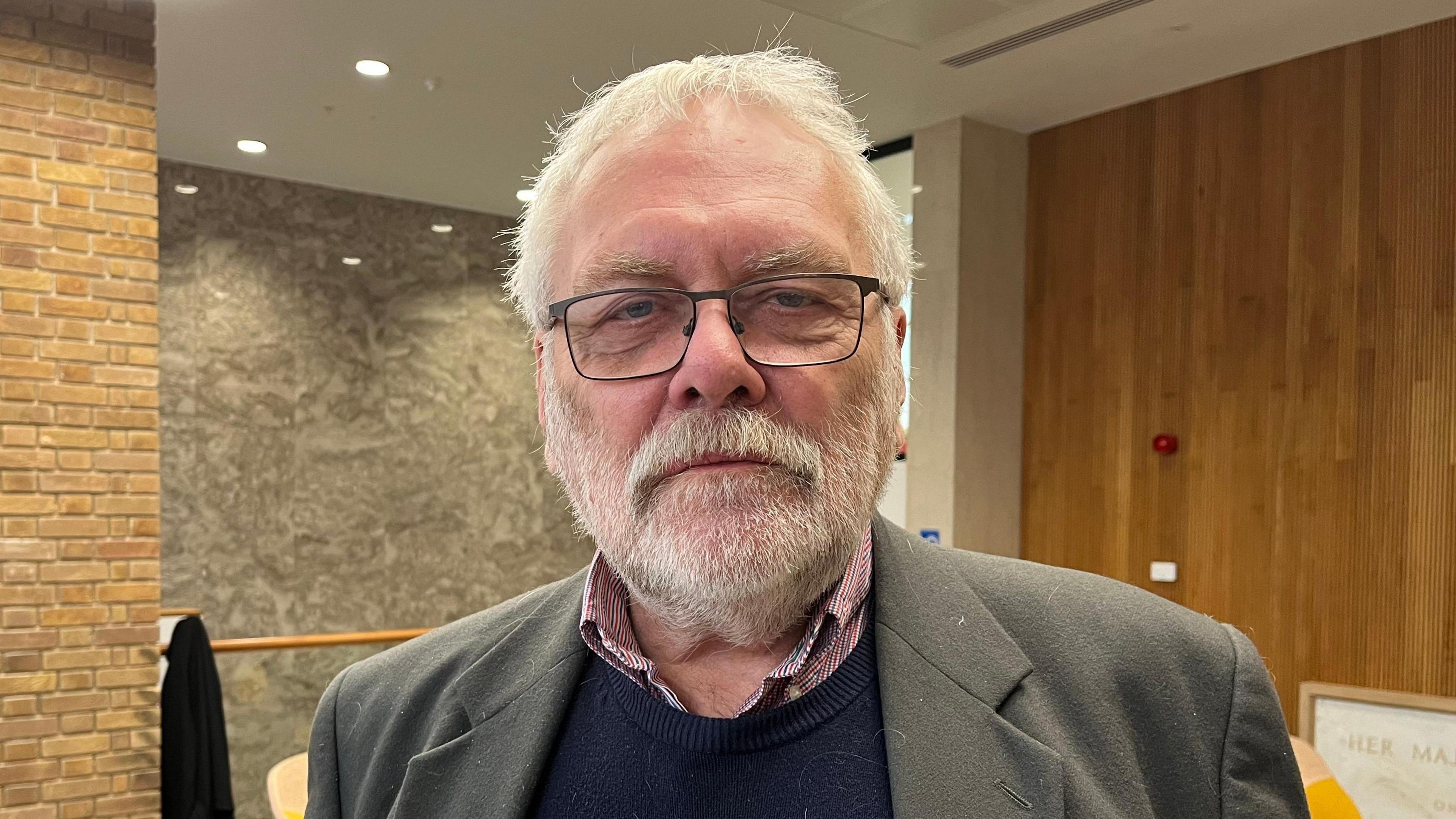 Steve Morphew is wearing a grey/light brown jacket over a navy jumper and red and white checked shirt.