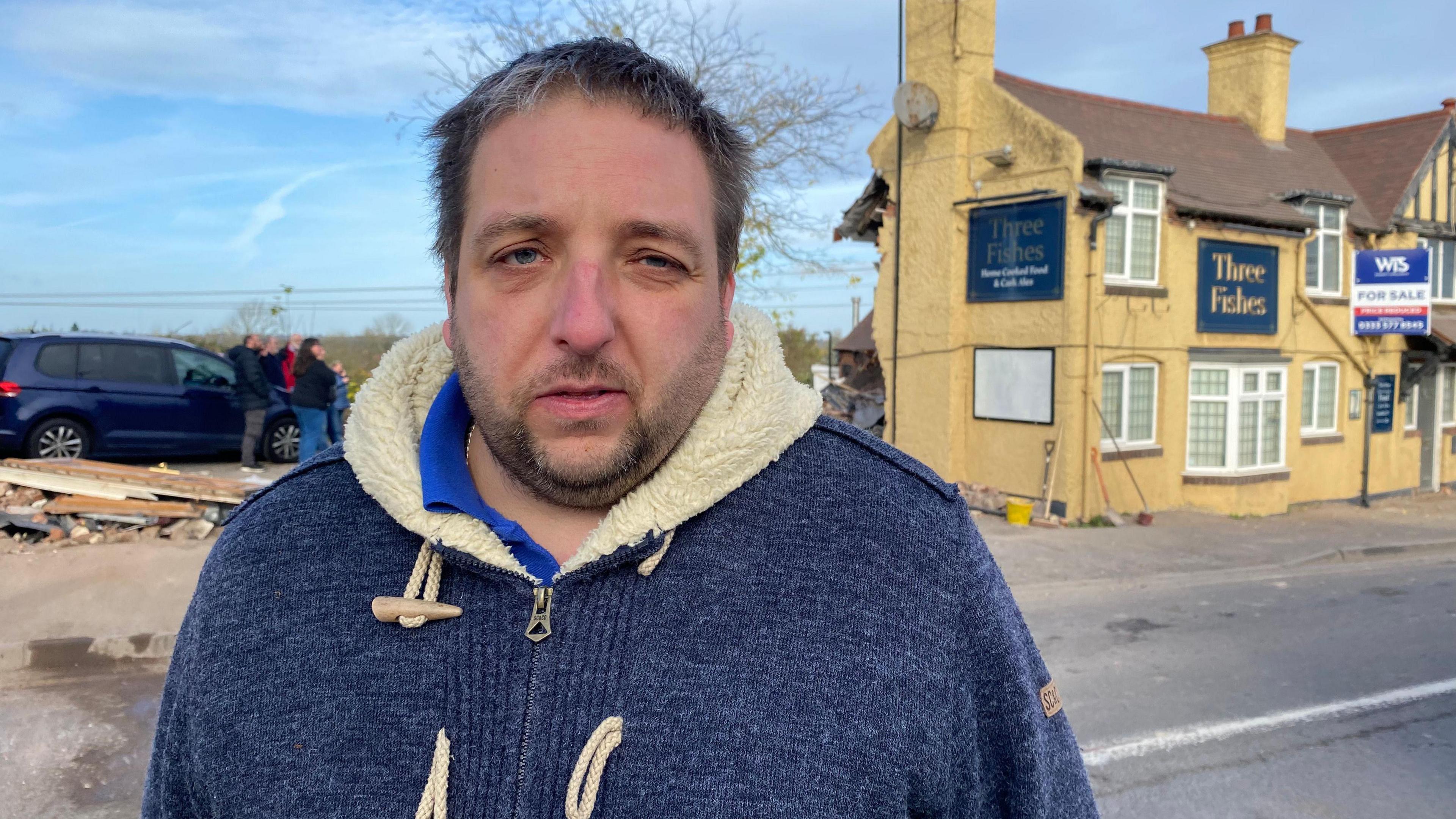 A man with a beard and a blue fleece standing in front of a beige-coloured pub with a blue sign saying Three Fishes