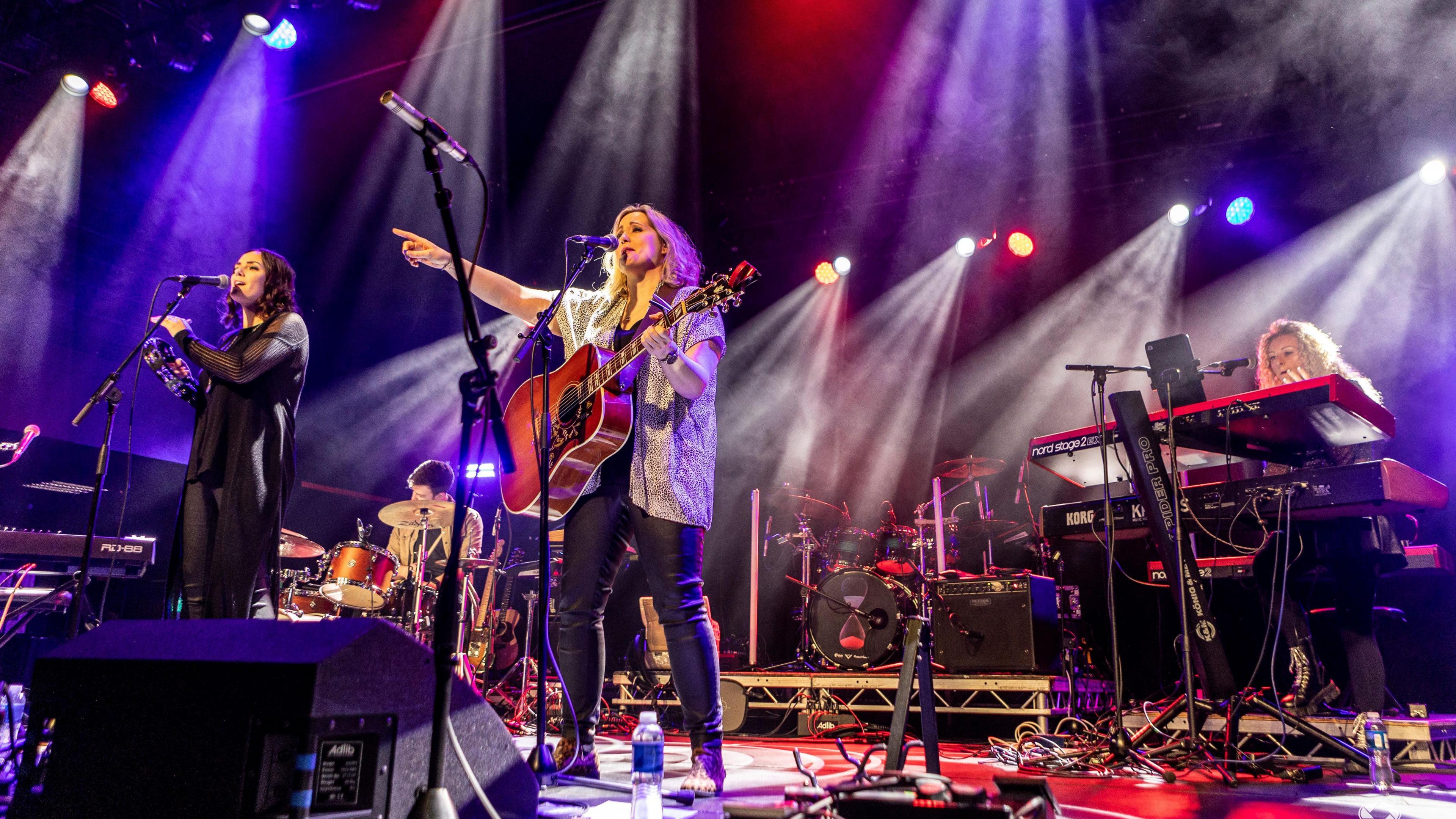 The Eves onstage - Marissa is singing on the left of the picture while Caroline is singing while holding a guitar. A keyboardist and drummer are behind them.