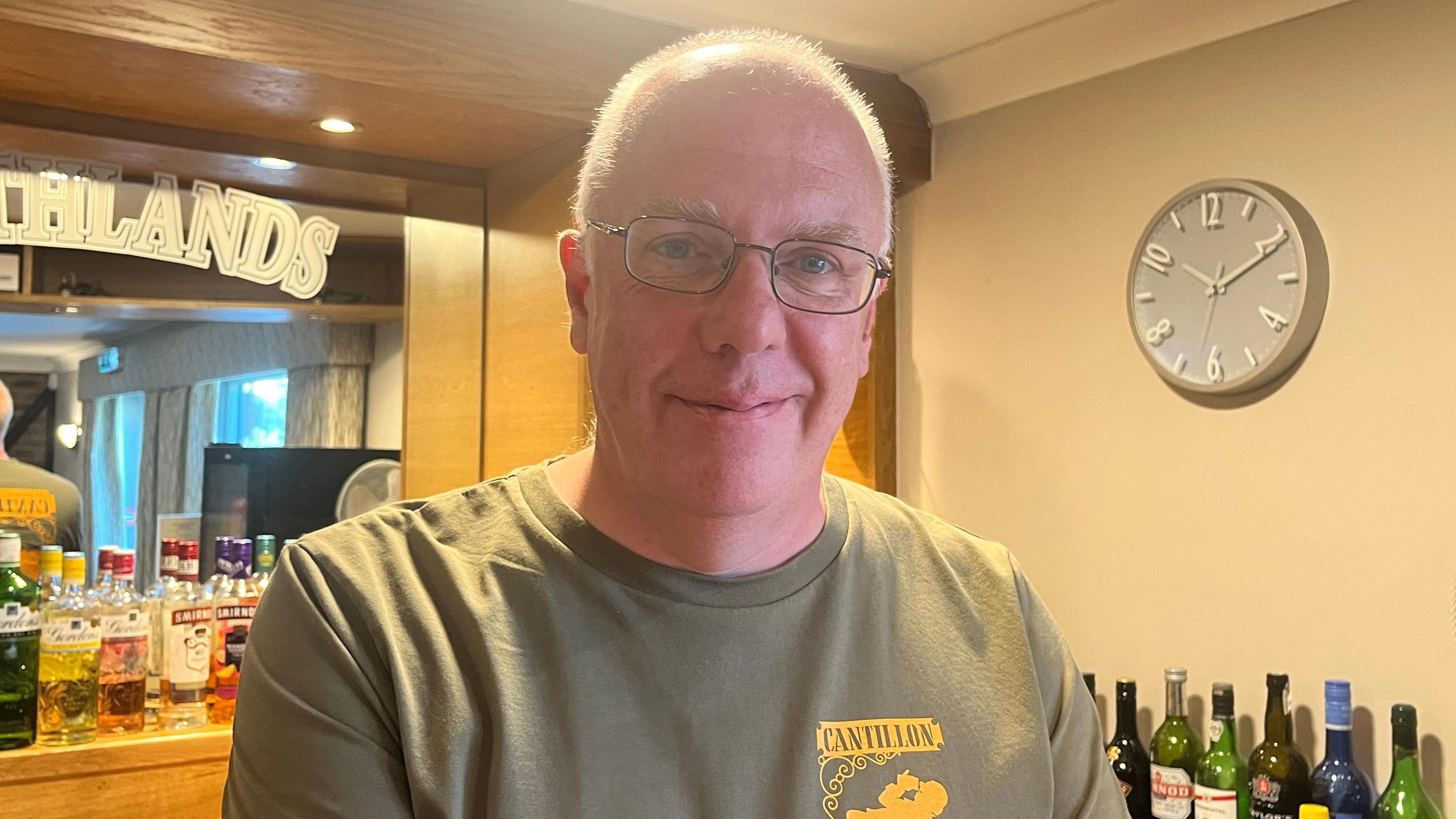 Paul Grainger has short white hair. He is wearing dark, slim framed glasses and is wearing a grey t-shirt with a yellow logo on the right. 