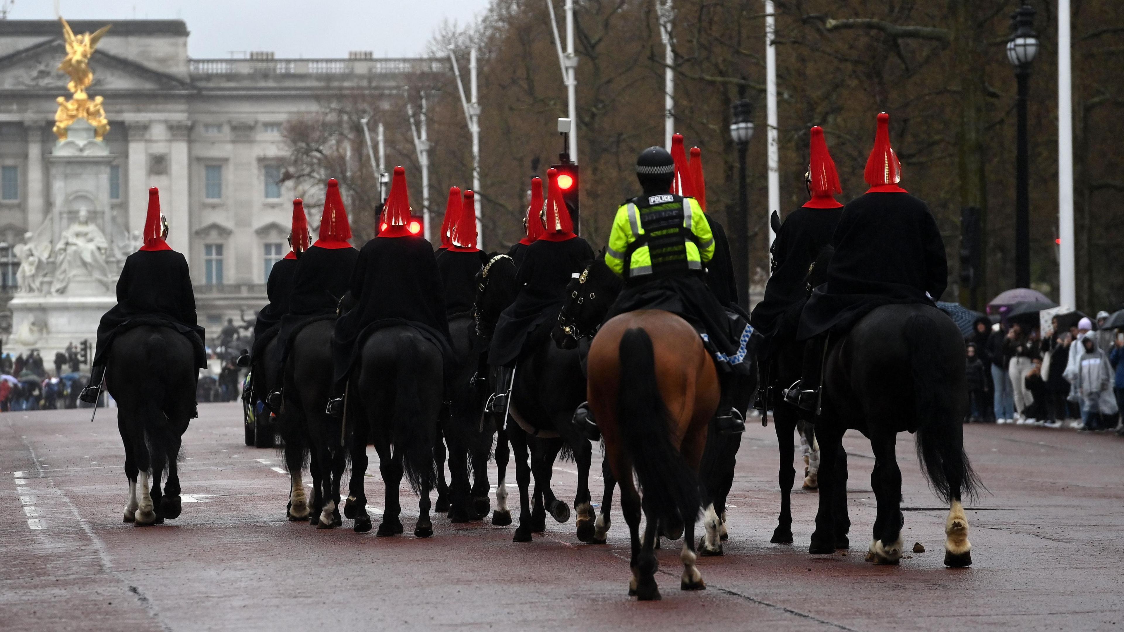 A group of soldiers, wearing red head pieces, are pictured on black horses along the Mall in central London. A police officer is shown in the centre of the picture on top a brown horse. Tourists and Buckingham Palace can be seen in the background. 