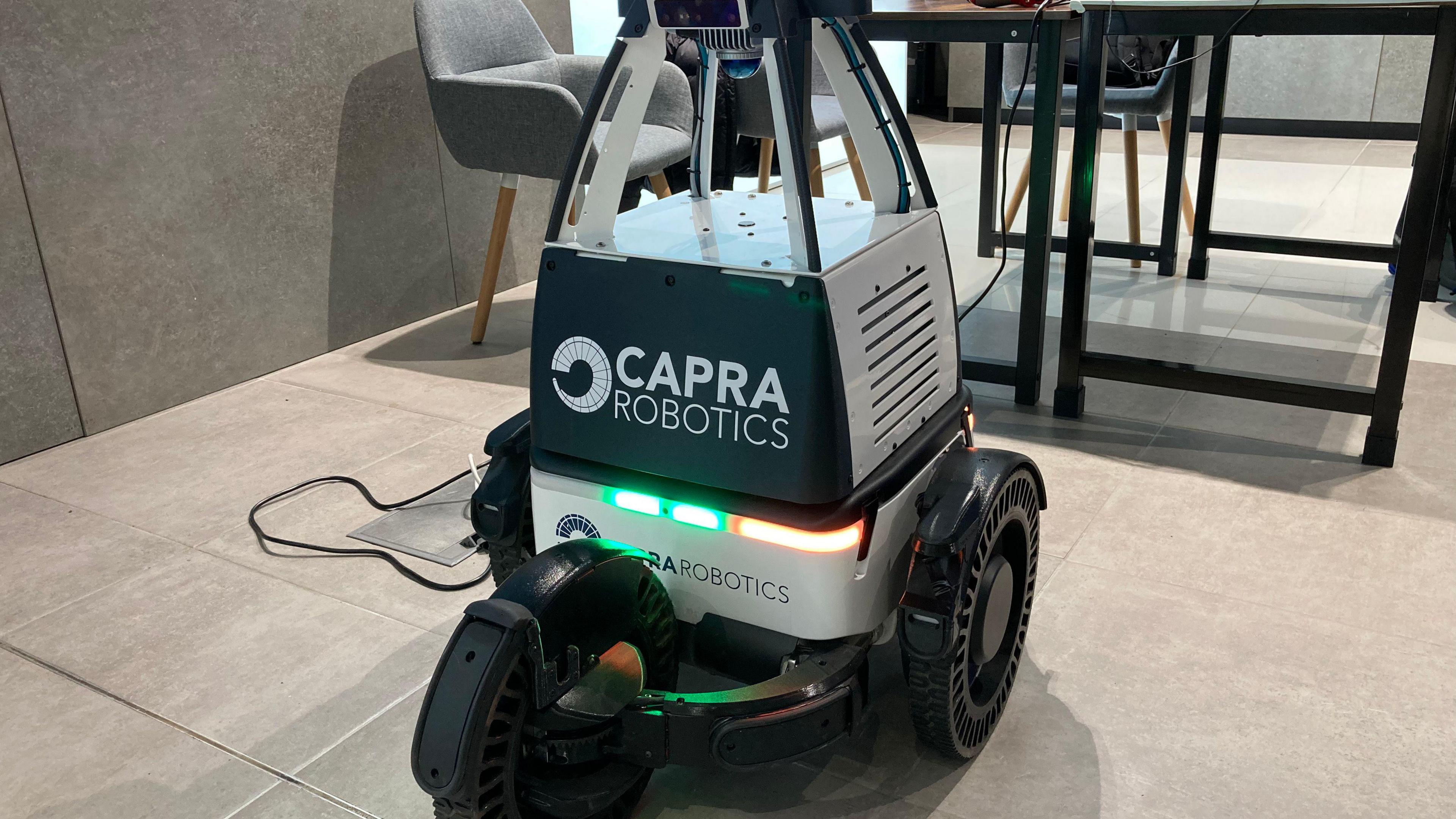 A robotic device is pictured on a tiled floor. It has three wheels and is black and silver. Printed on the front are the words "Capra Robotics"