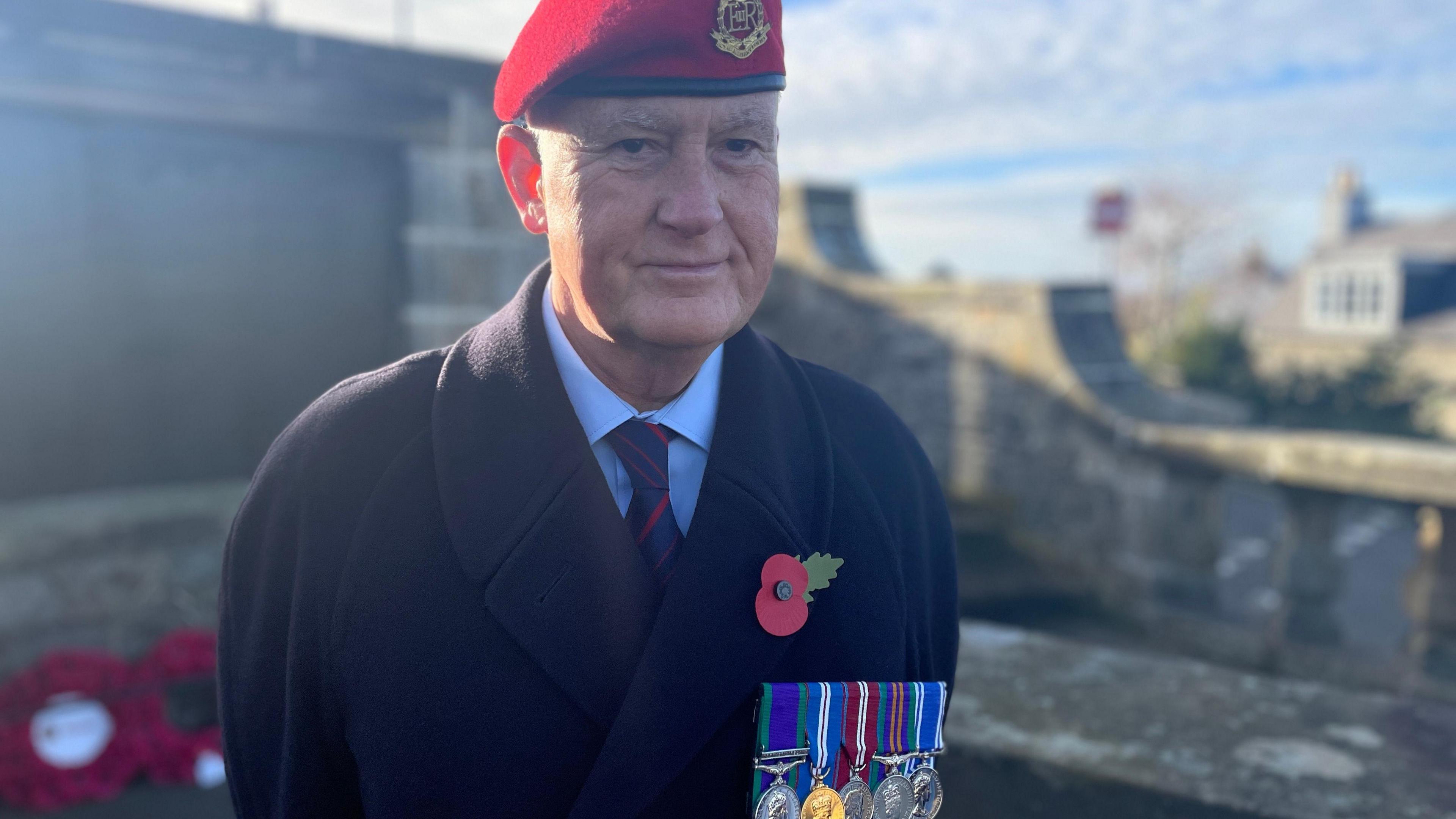 Mike Ashford-Smith, who has a poppy and five medals on striped ribbons on his navy woollen coat, wears a red Royal Military Police beret and stands in front of red poppy wreaths. 