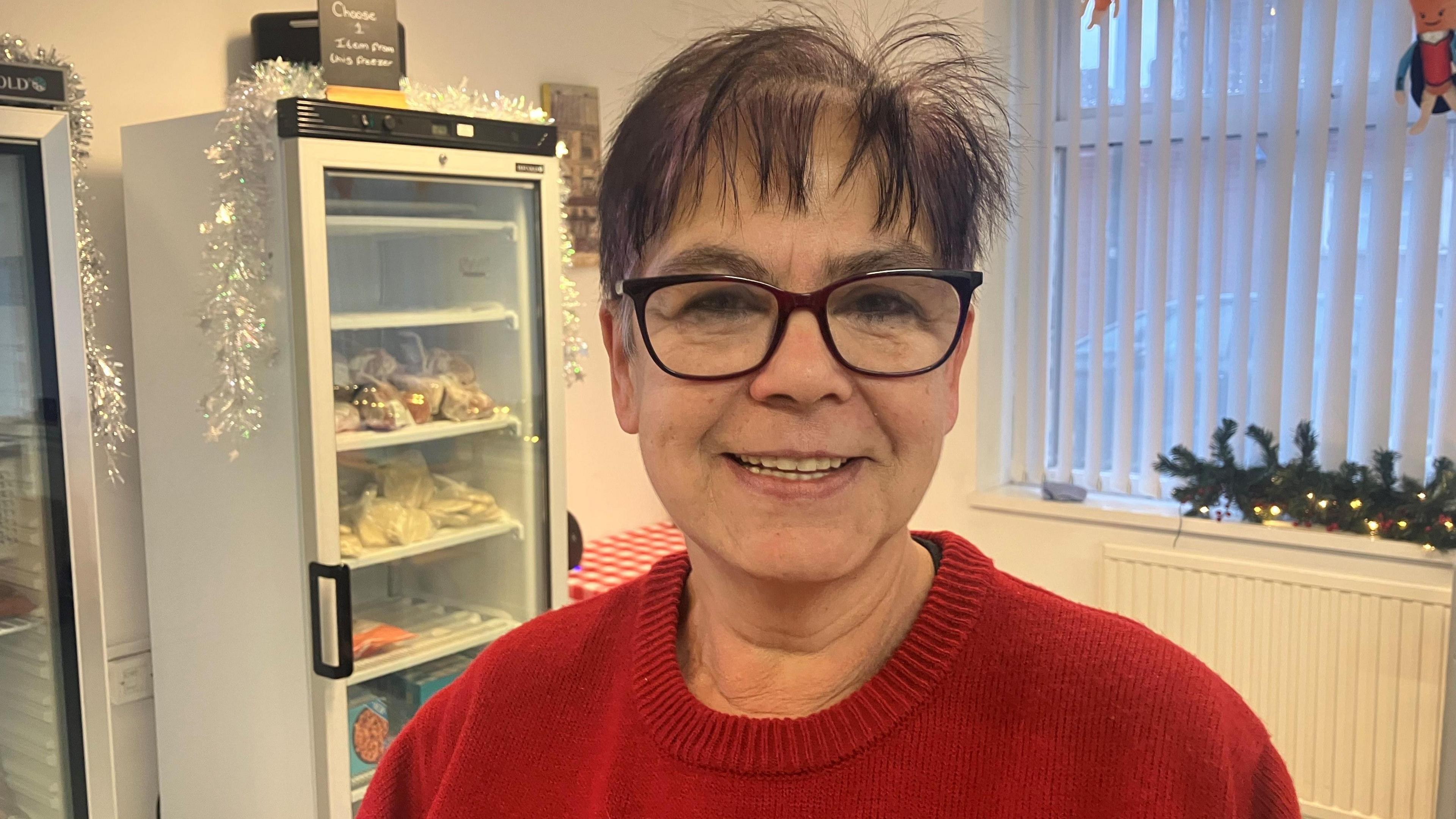 Photograph of Jazz Gough, who uses the Re:dish social supermarket in Stockport. With short, dark hair and wearing a round-necked red jumper, she is pictured in front of some fridges.