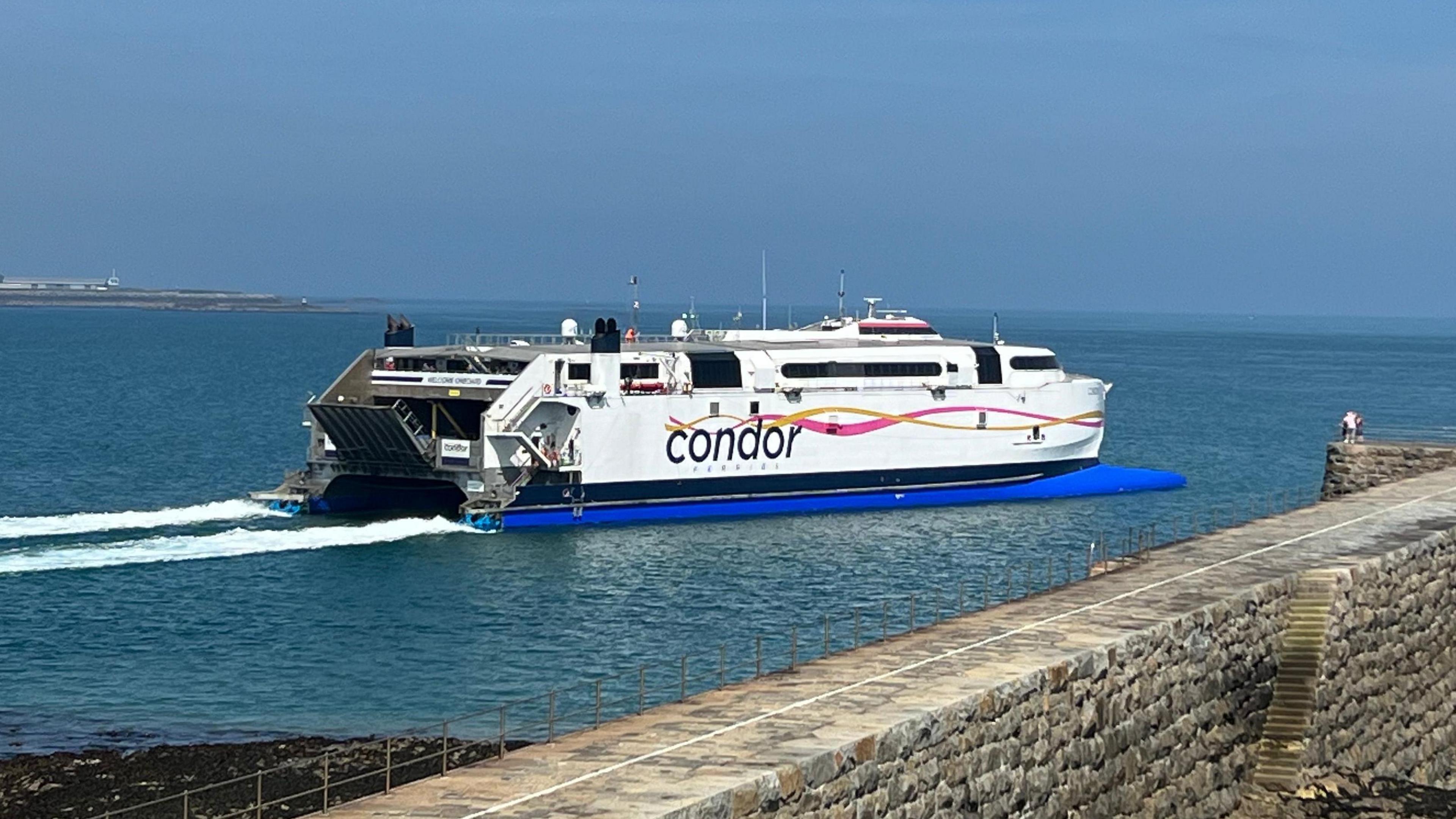 A Condor ferry heads out of a harbour which has a large grey brick wall.