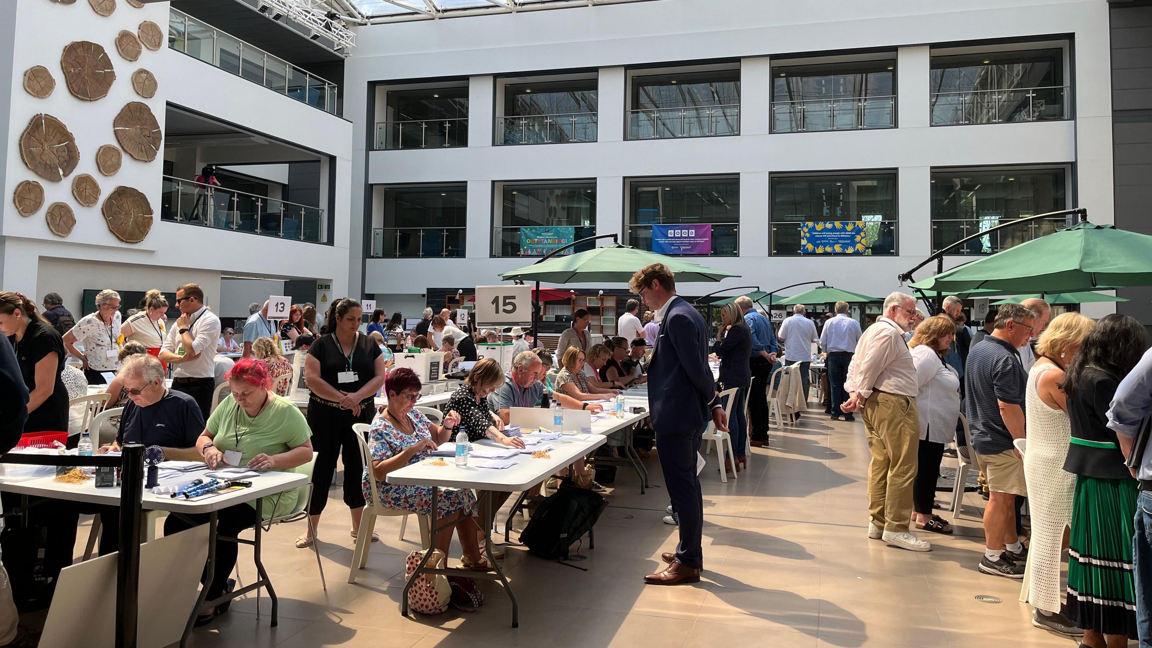 A crowd of people in an atrium, gathering around people sitting at trestle tables counting vote papers