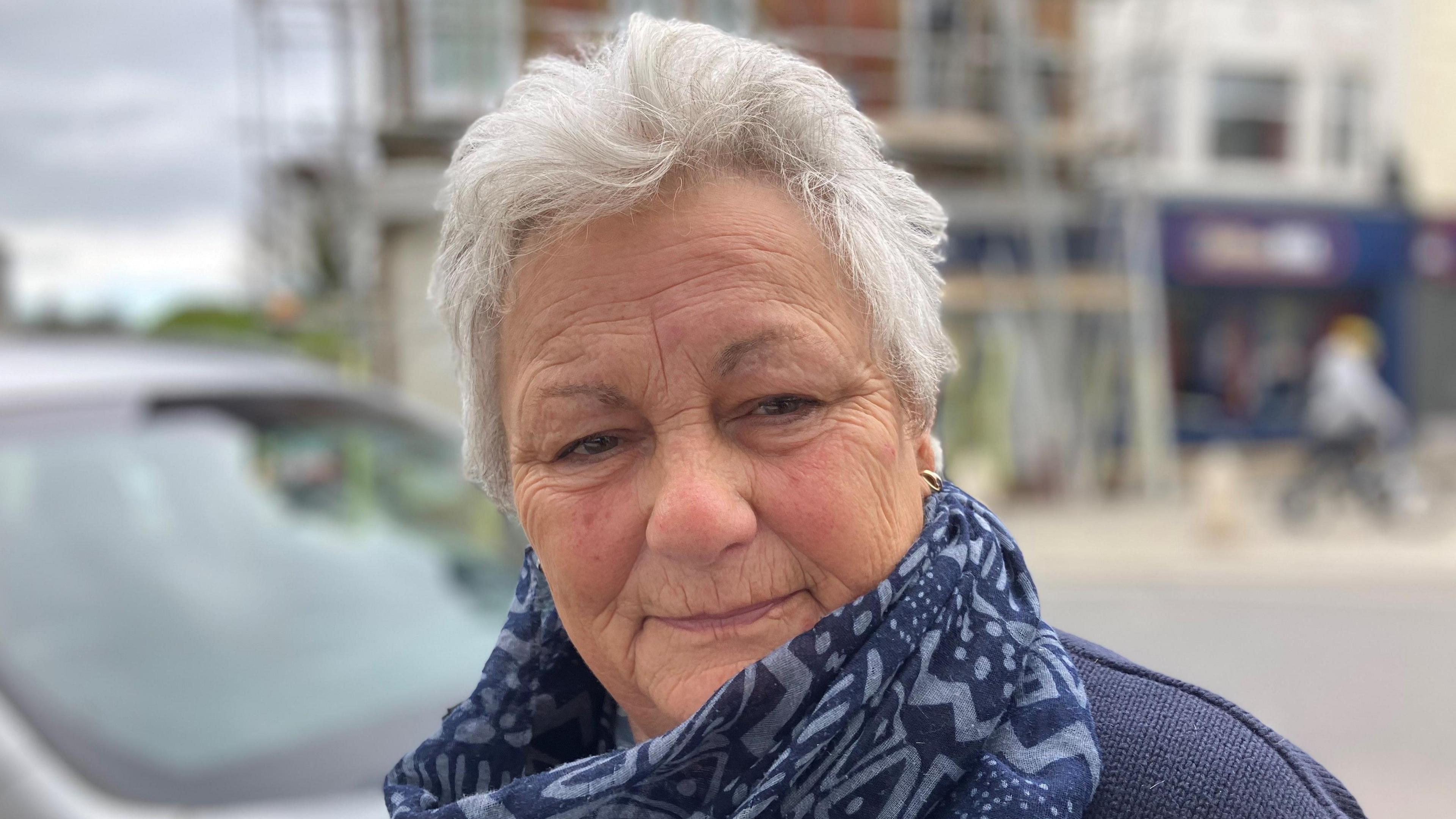 Mary Alford, who has white hair and a batik scarf. She is looking at the camera with a blurred picture of the centre of Launceston behind.