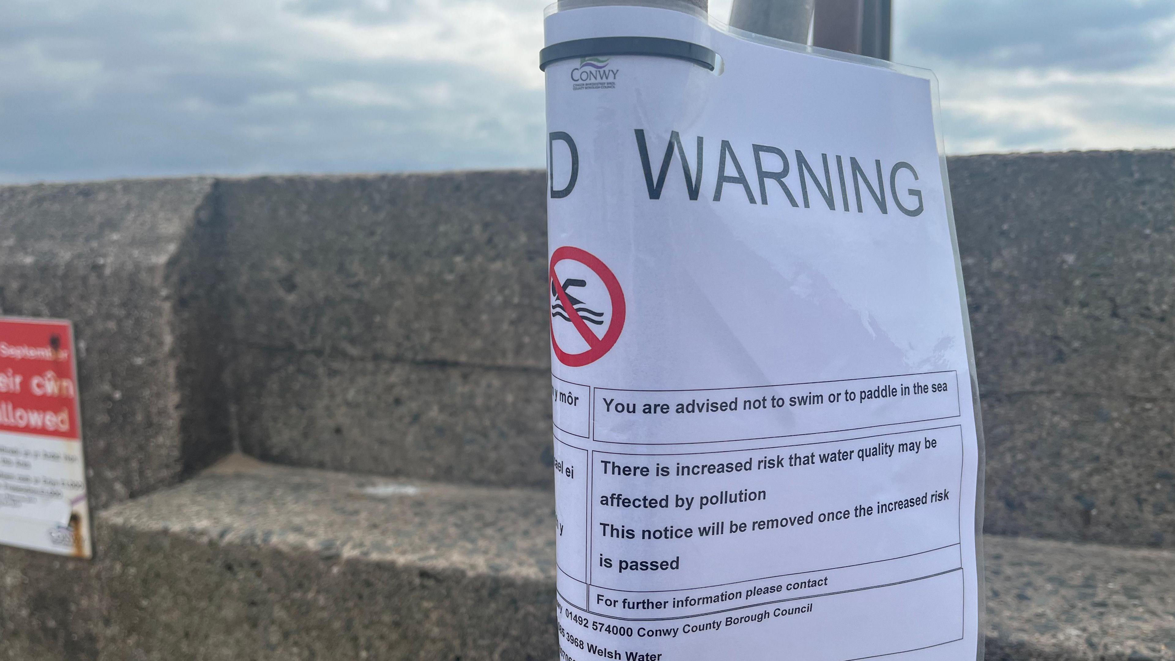 Paper sign taped to a pole and wrapped around it warning people not to swim or paddle in the sea. It reads: There is increased risk that water quality may be affected by pollution. The top of a concrete seawall on the promenade is visible behind