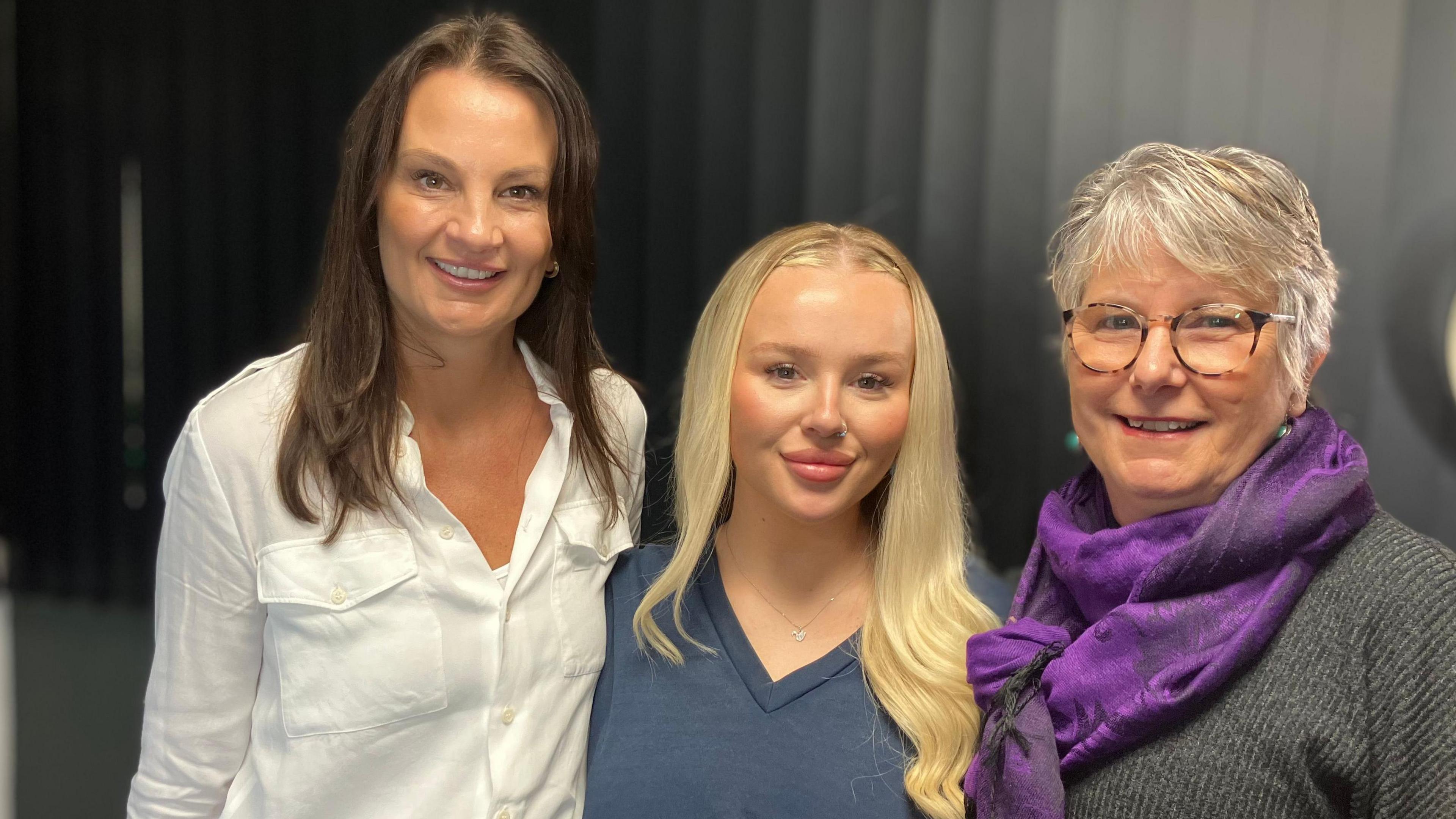 Left to right:  Sam, Bath and Anne. Sam has long brown hair and is wearing a white shirt. Beth has long blonde hair, a nose ring and is wearing a navy V-neck top. Anne has short grey hair and is wearing a grey top and is wearing a purple scarf 