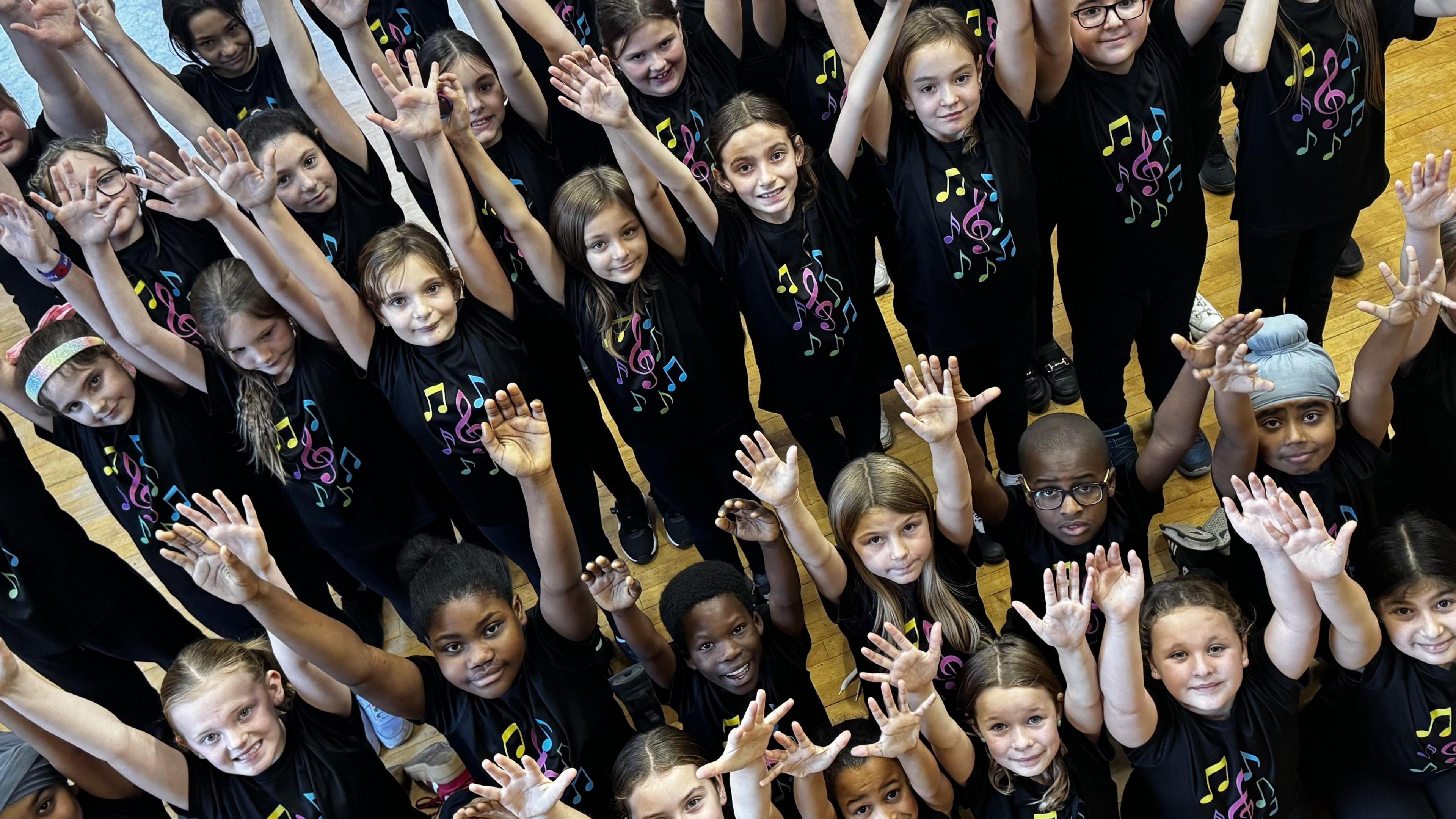 A group of small children in matching black t-shirts hold their hands in the air and smile up at the camera