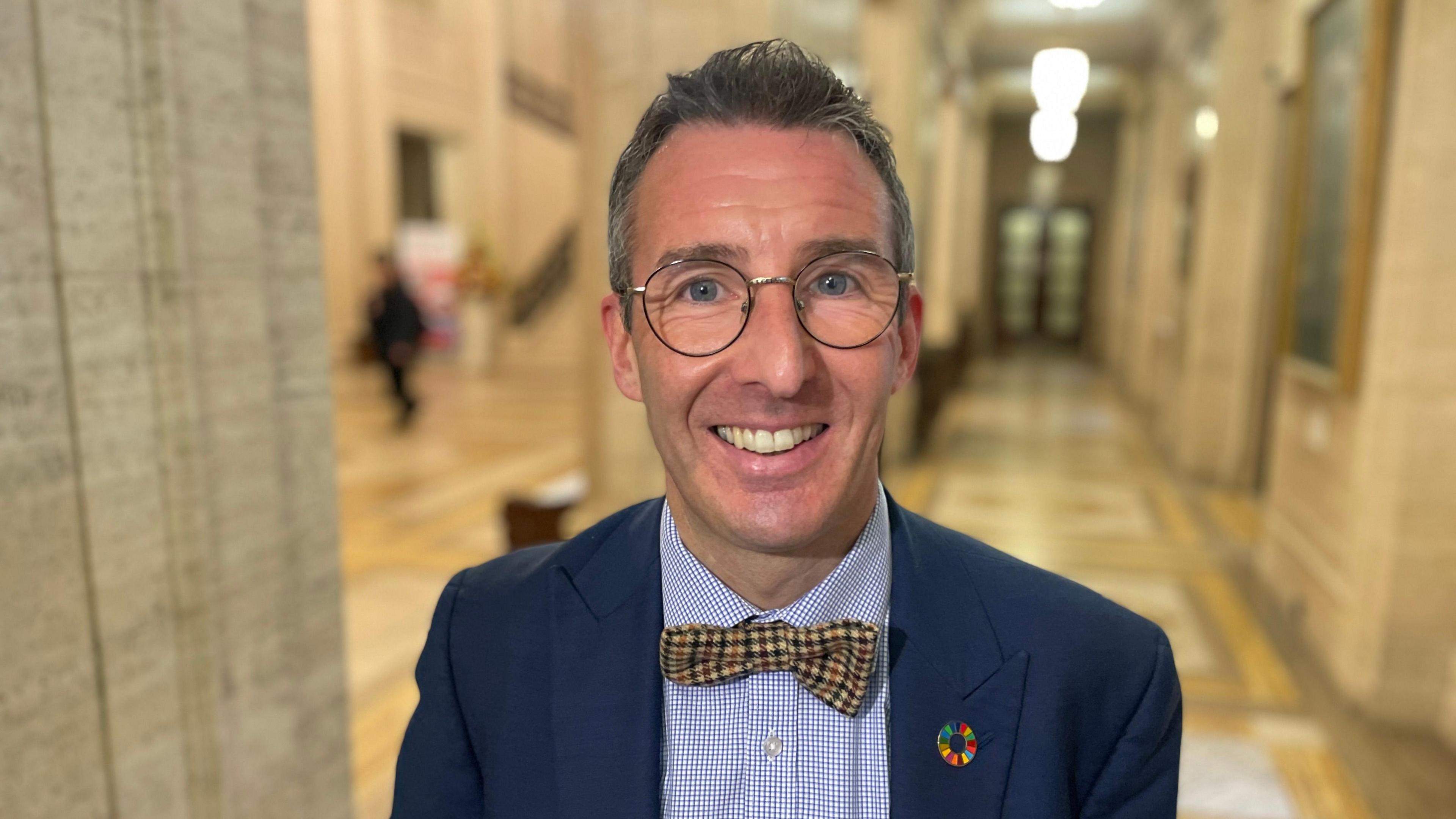 Andrew Muir. He has shirt grey-ish hair and blue eyes, wearing glasses, a blue blazer, blue check shirt and yellow check bow tie.