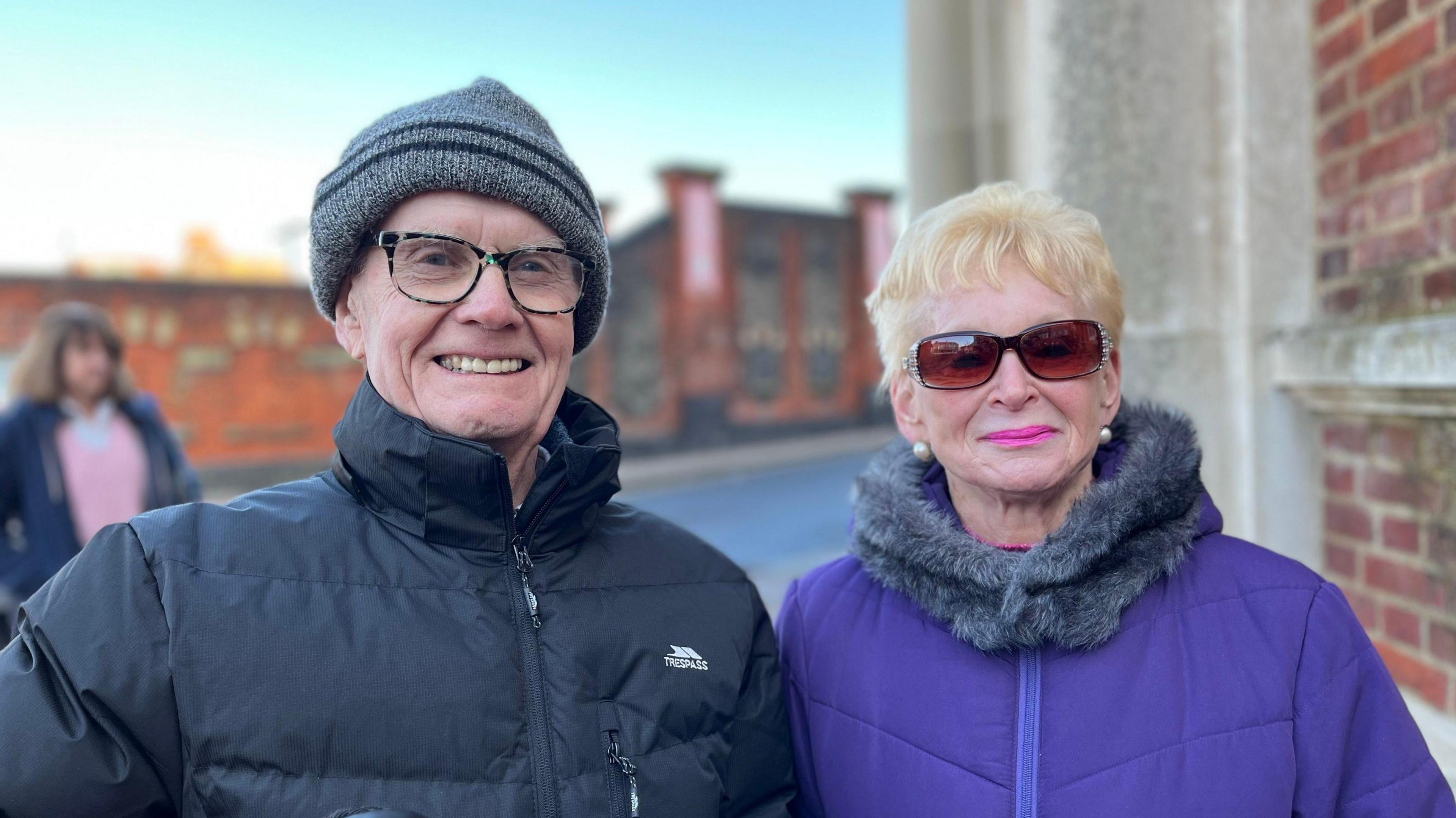 Laurence and Elaine Stringer standing outside the red bricked post office smiling at the camera. Laurence is wearing a black winter jacket and holding a daschund, Elaine is wearing a purple winter jacket and sunglasses and has short blonde hair