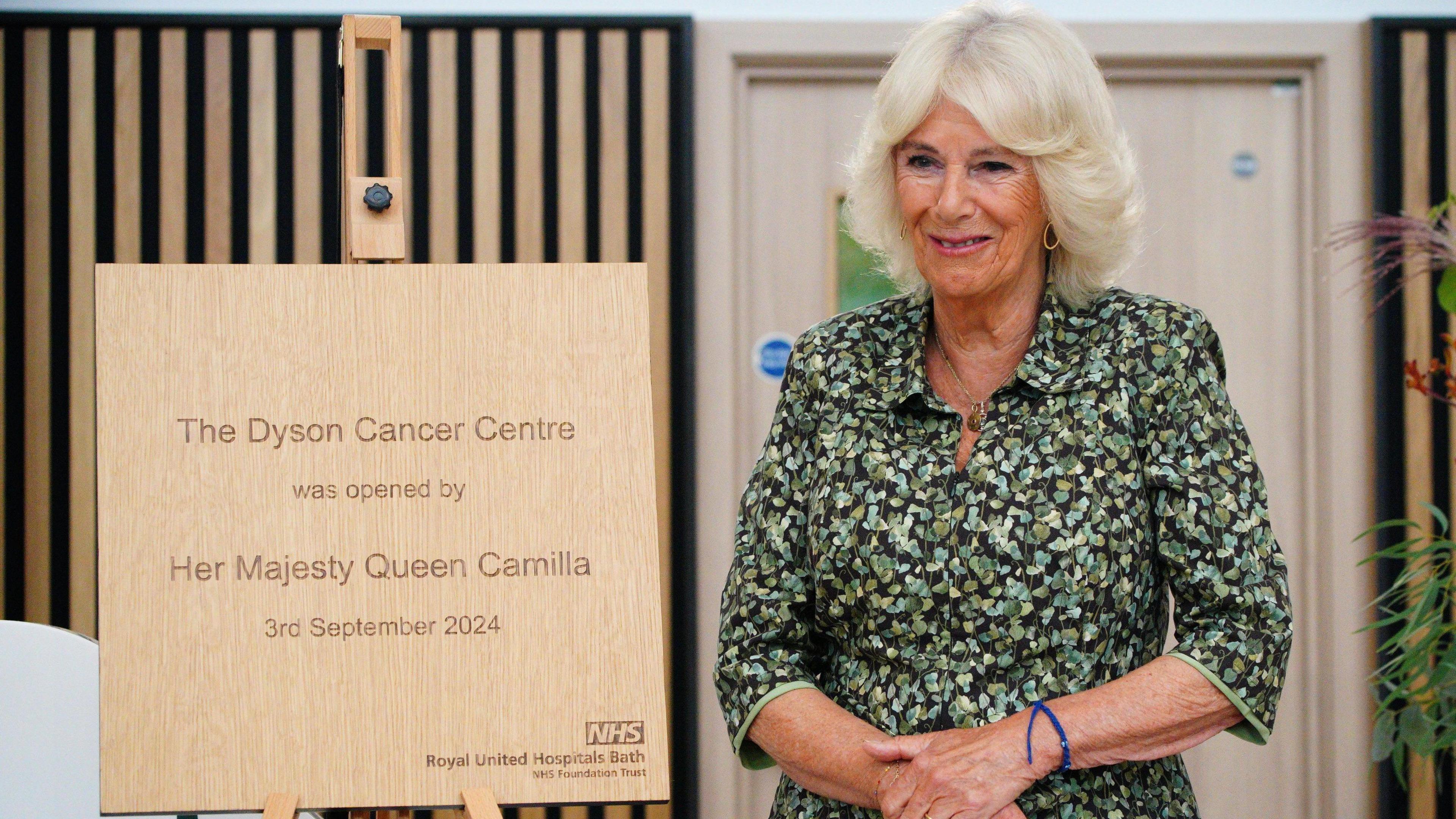 Queen Camilla standing next to a plaque on an easel, smiling