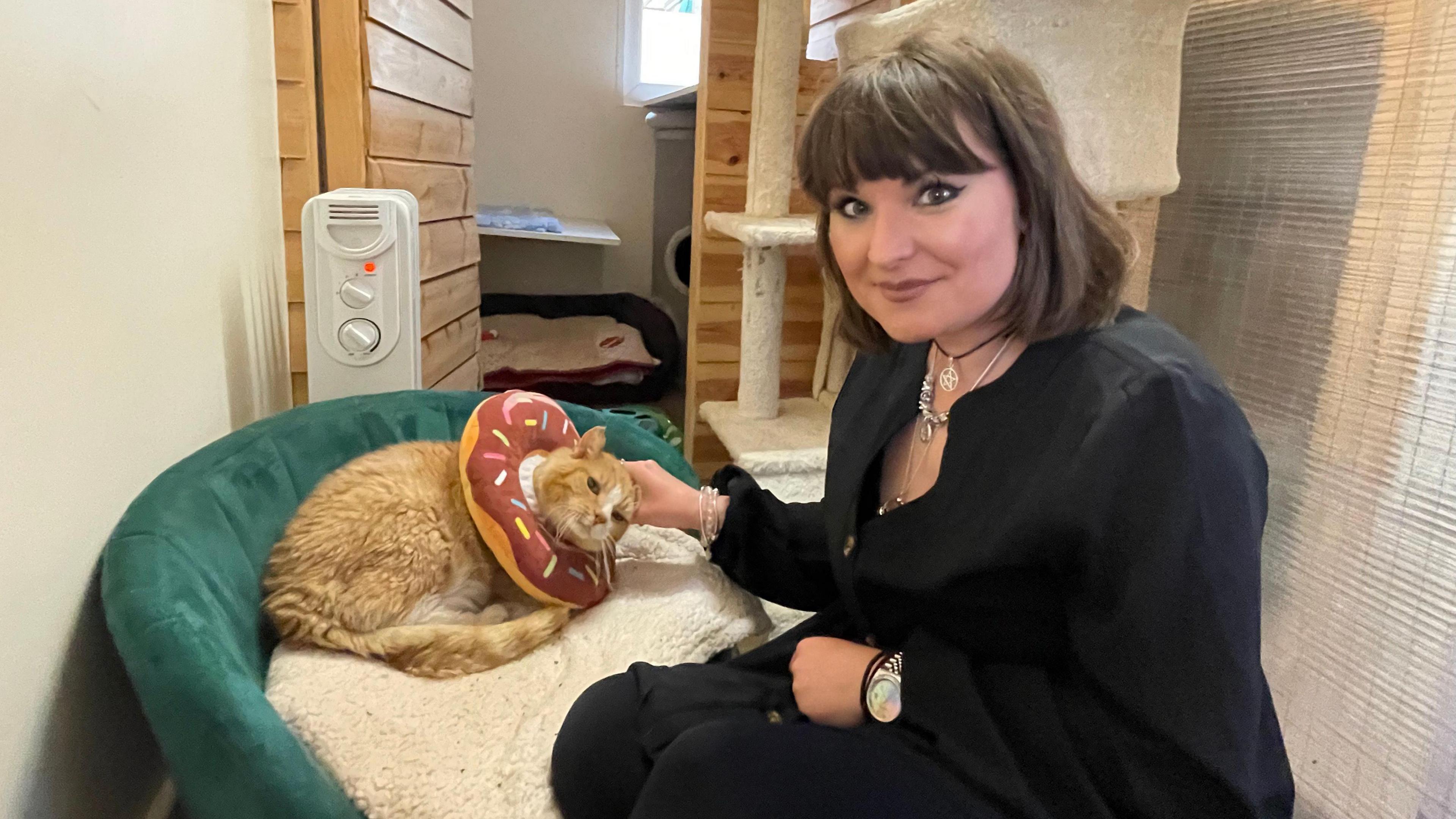 Fosterer Jemma crouches next to an elderly ginger cat, Murphy and strokes his head. He is wearing a soft doughnut neck brace.