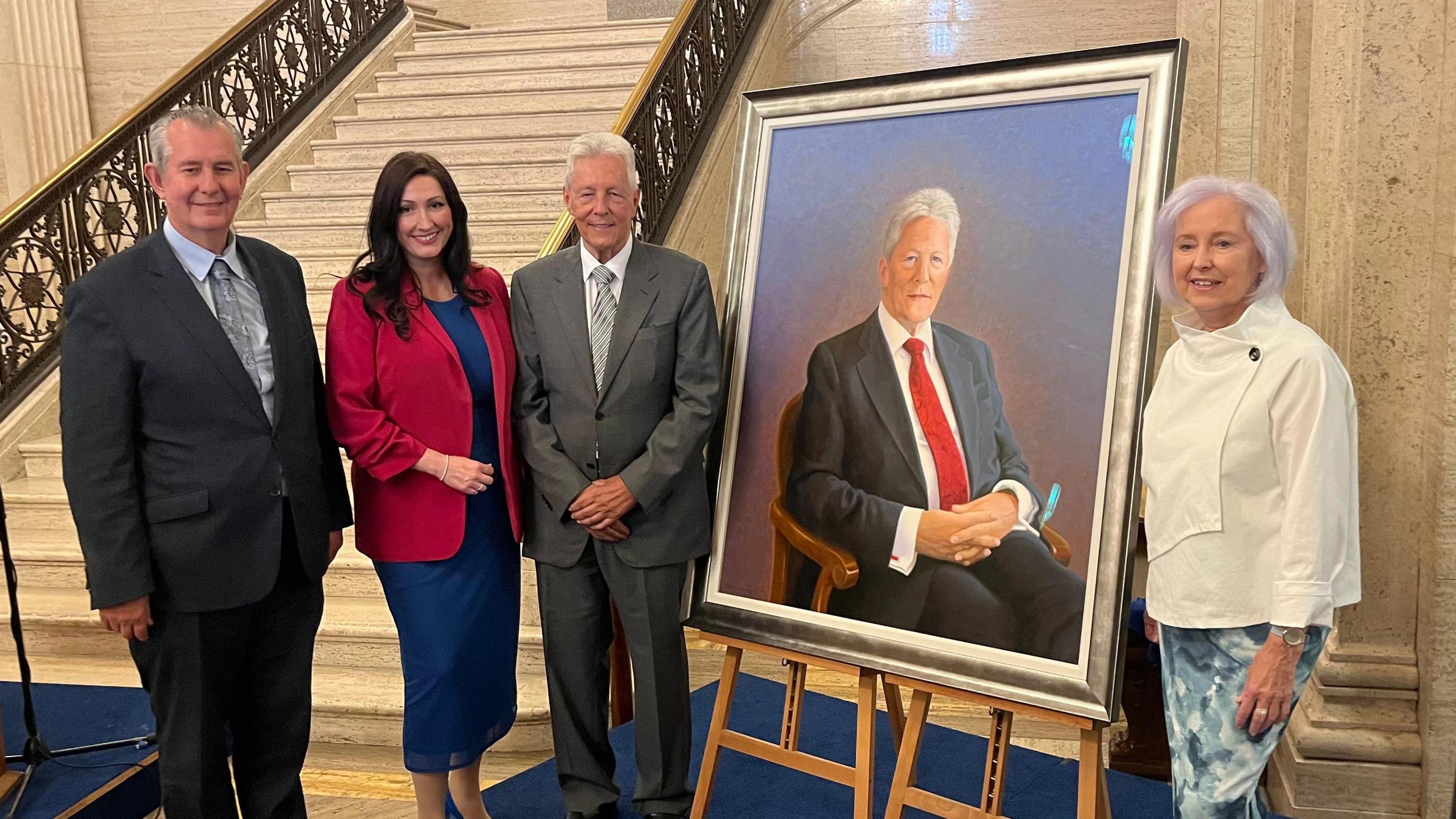 Edwin Poots, Emma Little-Pengelly, Peter Robinson and Carol Graham standing by the portrait of Peter Robinson at Stormont. They are standing beside a cream marble staircase. Poots and Robinson are both in suits, while Little-Pengelly is in a blue dress with a red blazer. Carol Graham is standing next to her portrait of a seated Peter Robinson, which is on display on a wooden easel. She is wearing a cream blazer and patterned skirt,