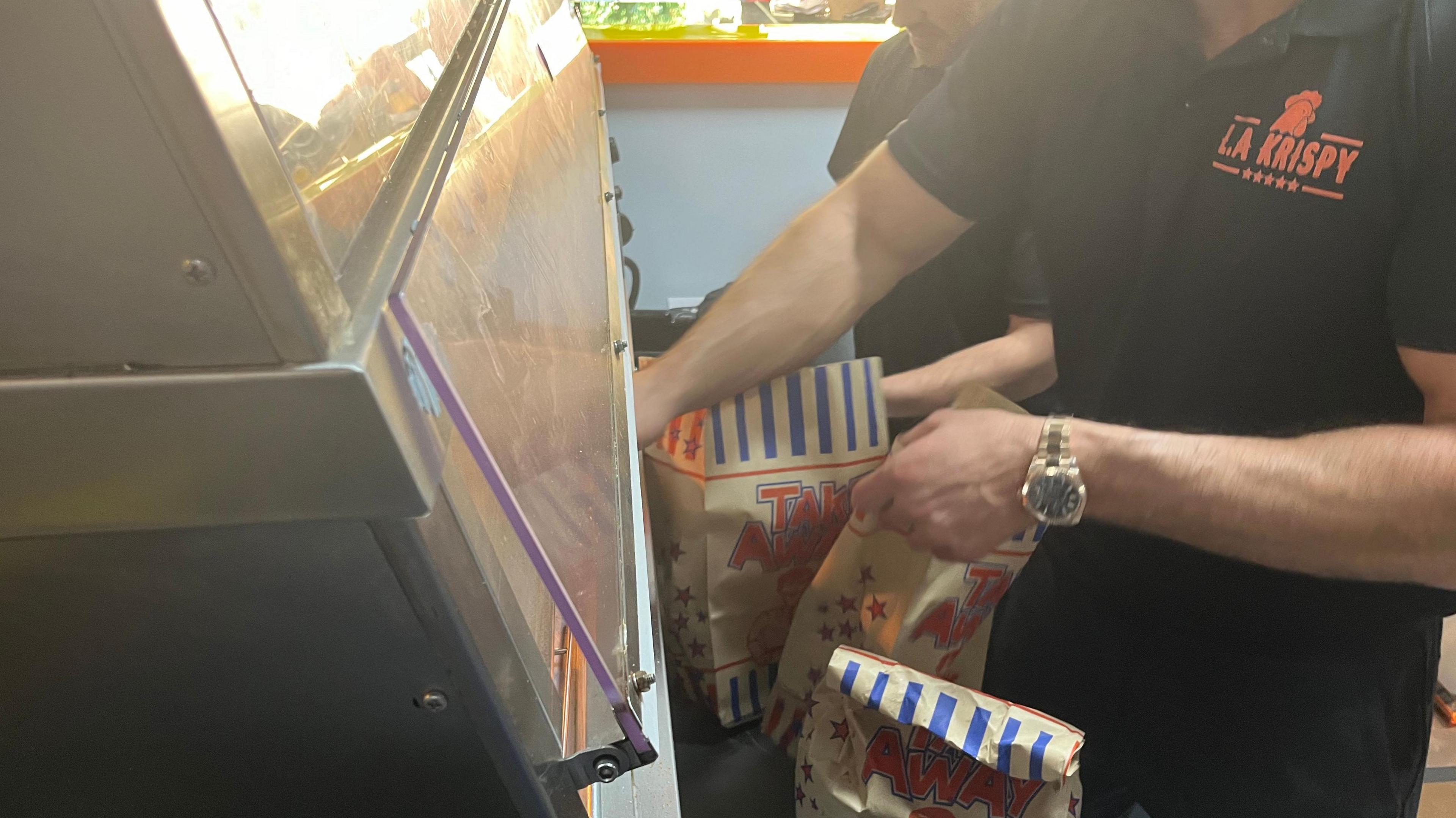 LA Krispy takeaway staff wearing black shirts with a logo in orange on them are packing orders into paper bags