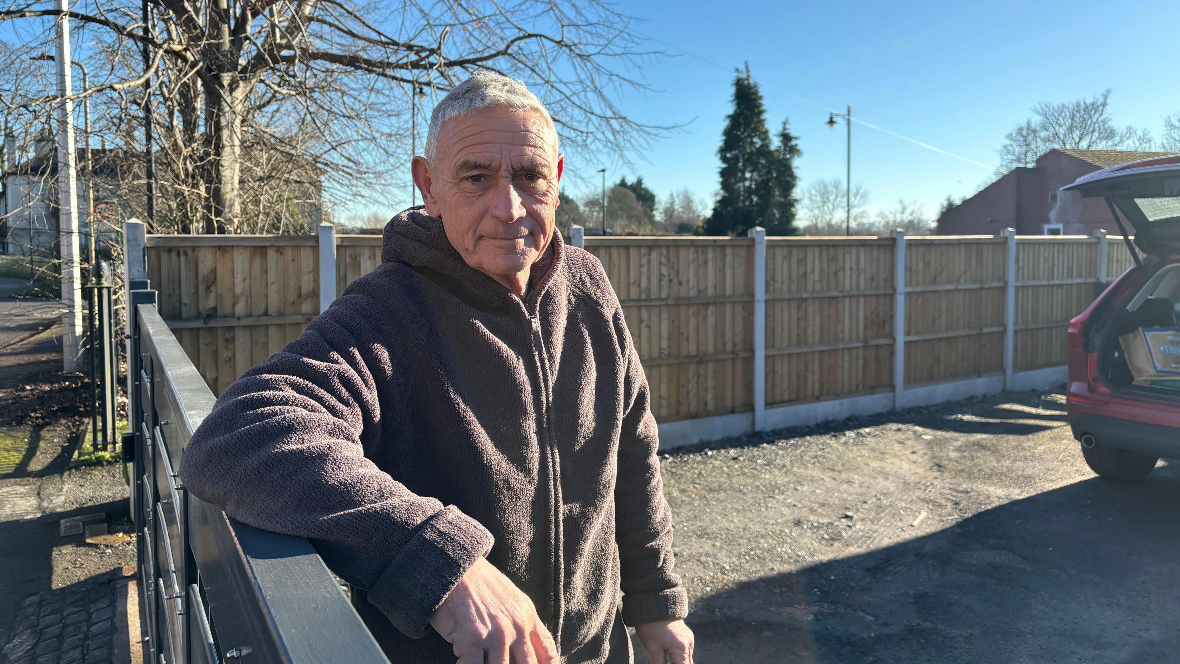 A man wearing a grey hoodie with his arm leaning on a fence. He is stood in a drive with a car boot open